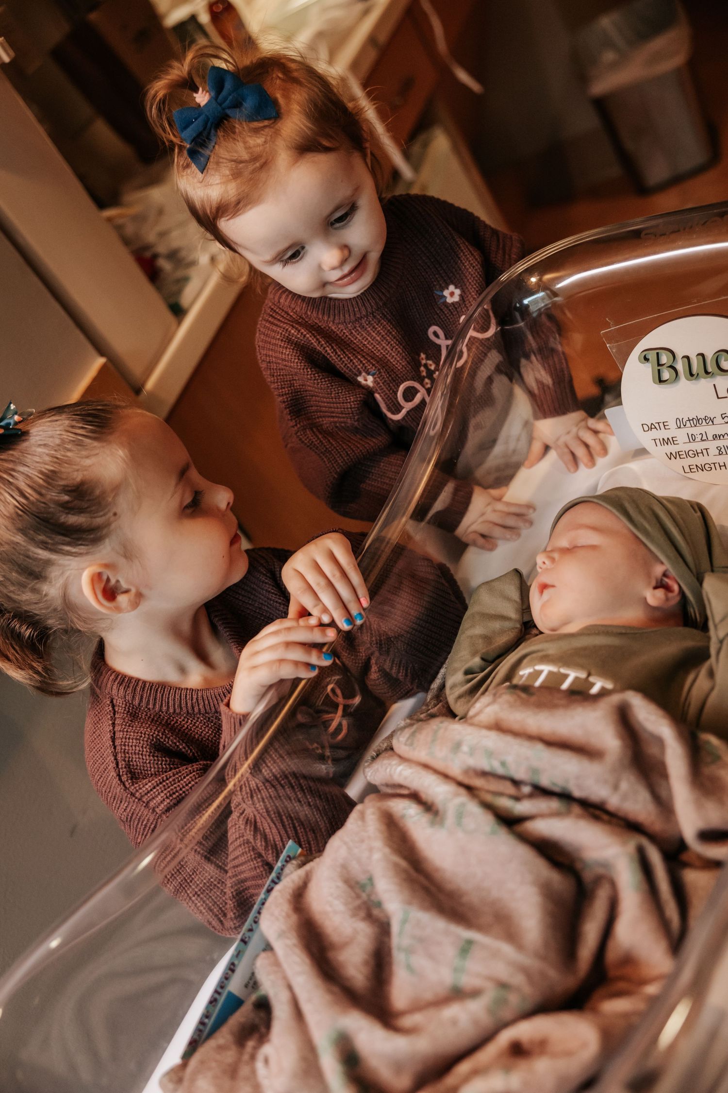Two young sisters in matching burgundy sweaters look at their sleeping newborn baby brother in a clear hospital bassinet.