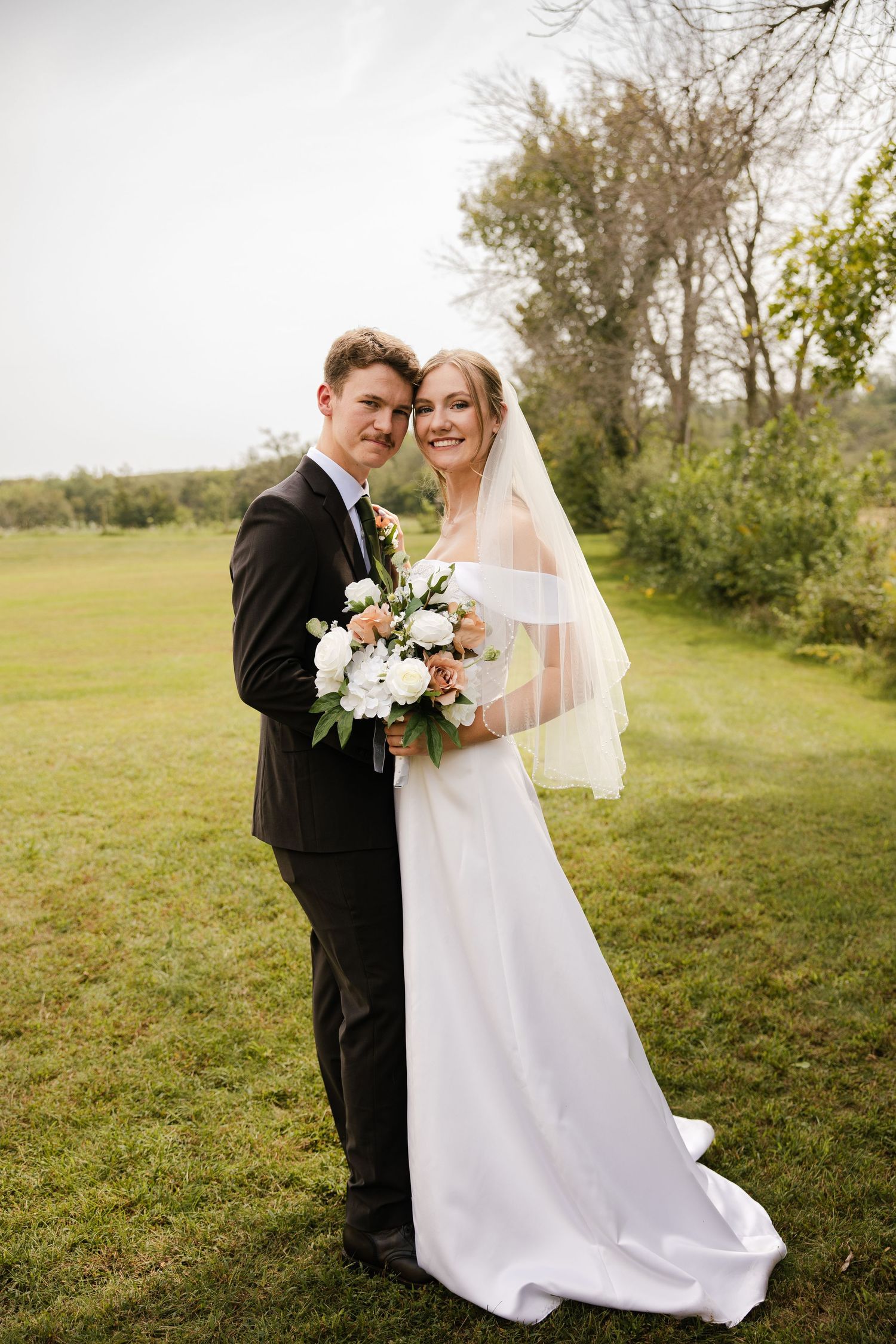 Smiling bride in a white gown and veil poses with her groom holding a rose bouquet in a grassy meadow.