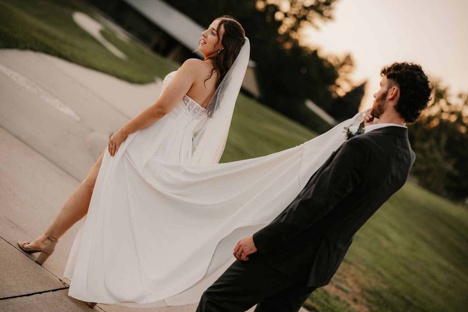 Bride in a flowing white gown and veil leans back beside the groom outdoors at golden hour sunset.