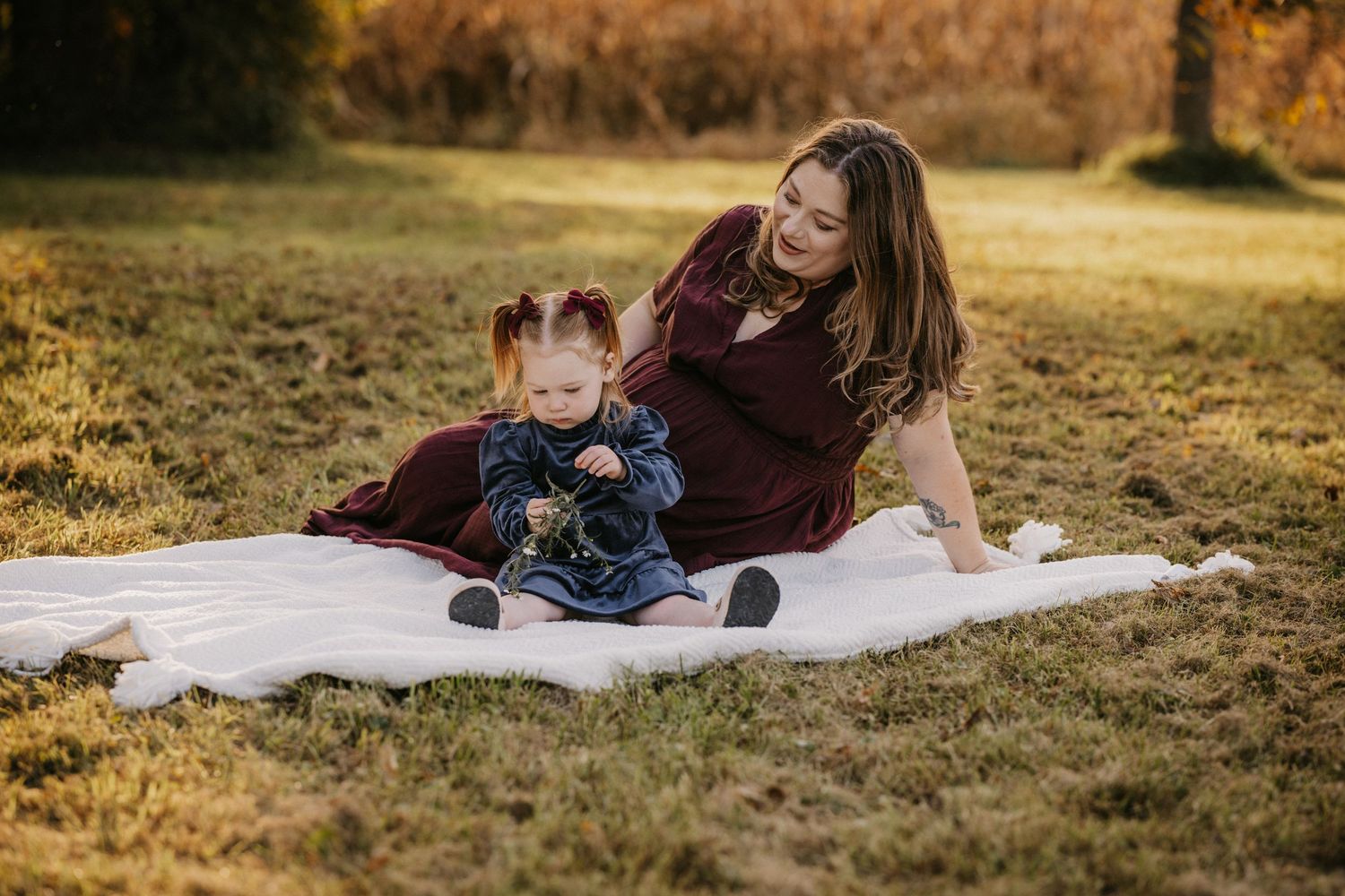 Smiling mother in a burgundy dress and her toddler daughter sit together on a white blanket in a sunlit grassy field.