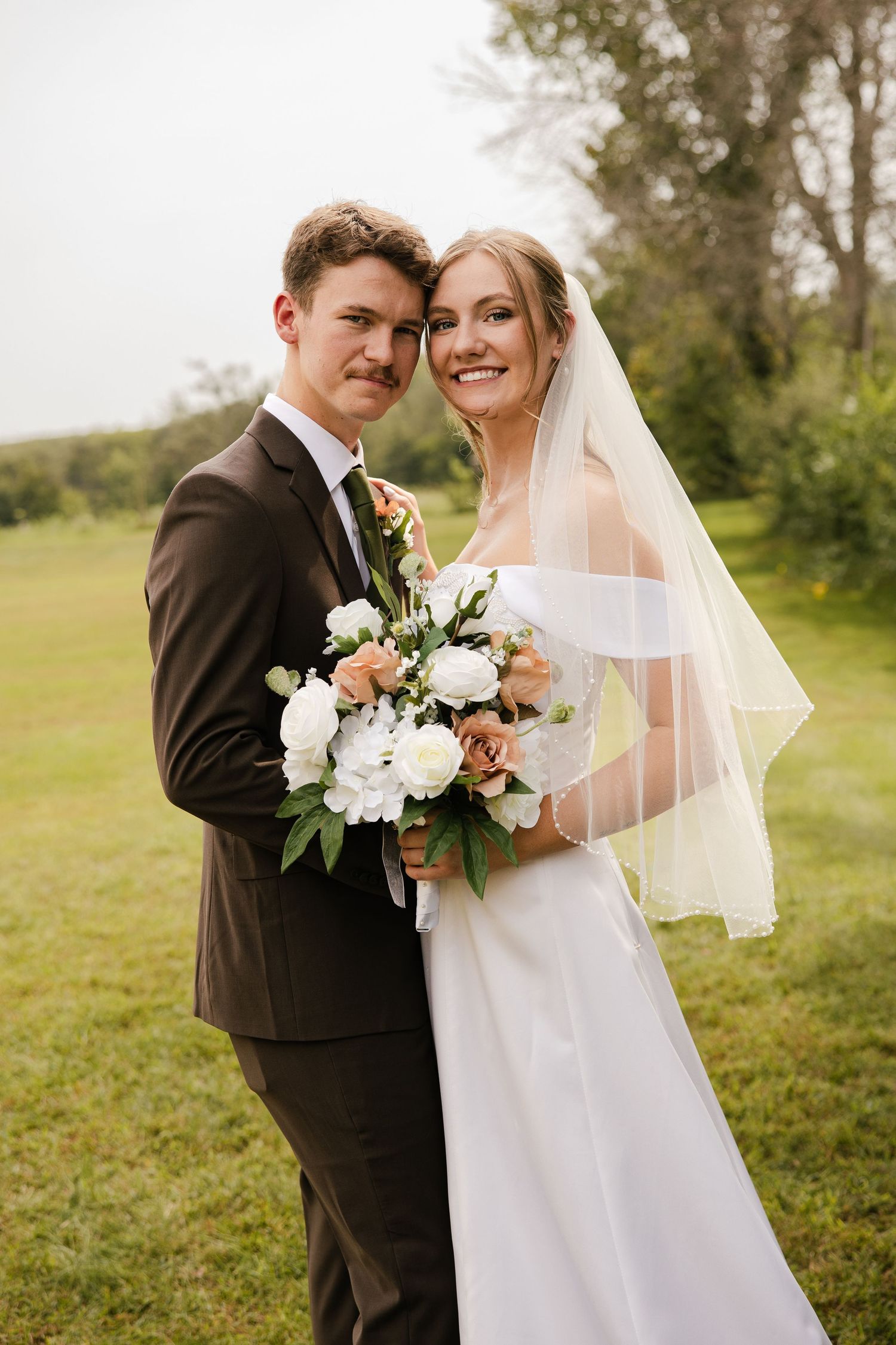 Happy newlyweds in a brown suit and white off-shoulder gown pose with a white and blush bouquet outdoors.