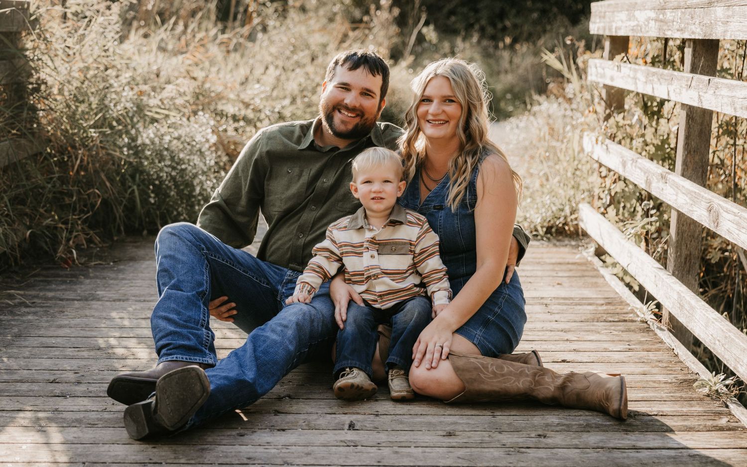 A smiling family of three sits together on a wooden bridge, with a bearded dad, a mom in cowboy boots, and a toddler between them.