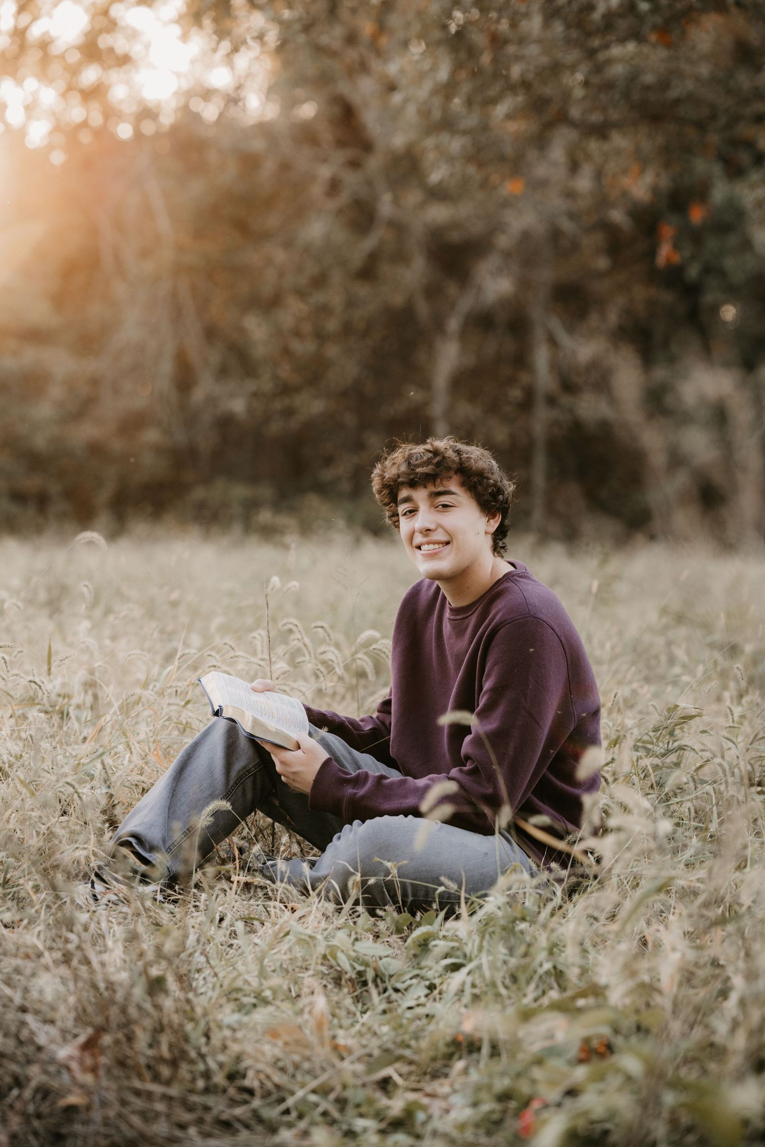 Smiling young man in a maroon sweatshirt sits in a field of tall grass holding an open book in warm golden evening light.