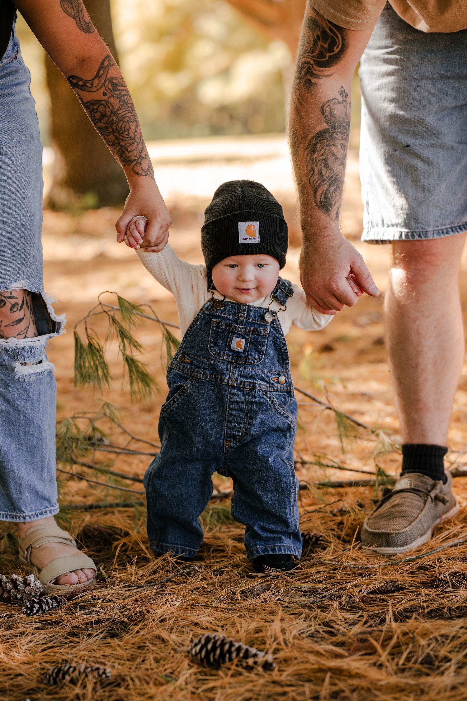 Baby in a black beanie and denim overalls stands on pine needles while two tattooed parents hold both hands in a woodland family photo.