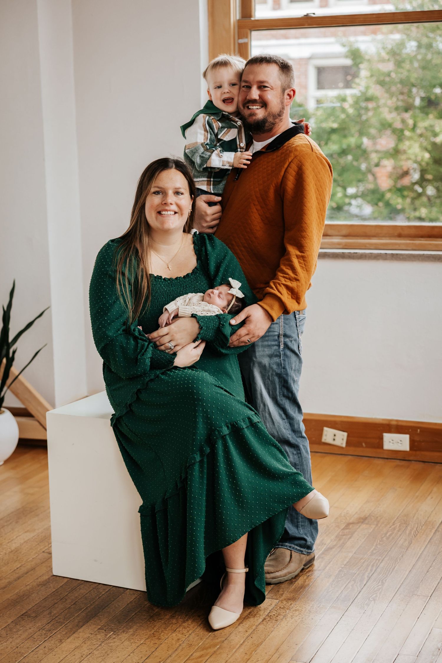 A mother in a green dress holds a newborn while a bearded father lifts a toddler beside a bright window in a family portrait.