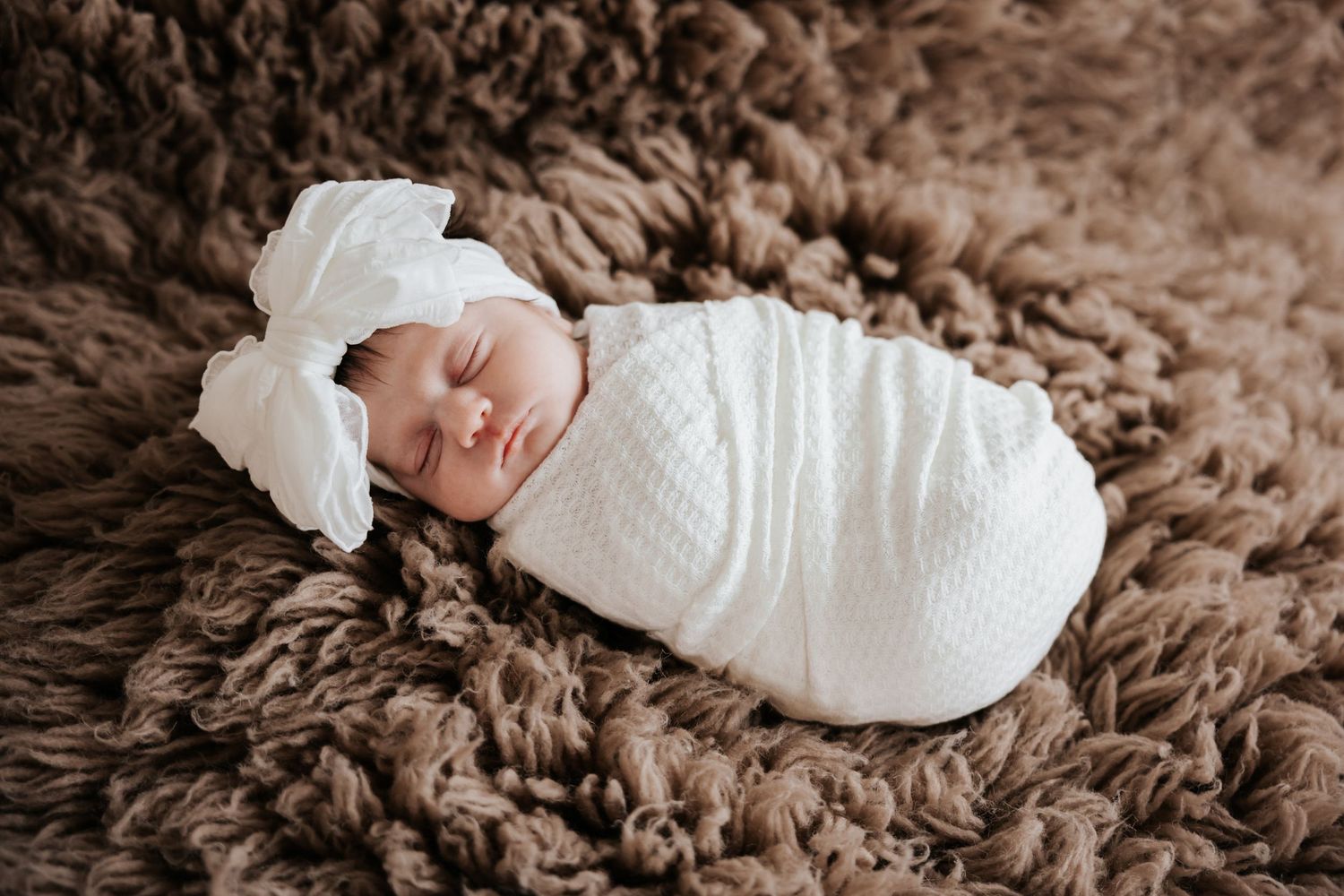 Sleeping newborn baby wrapped in a white knit swaddle and a large white bow headband rests on a brown shaggy rug.