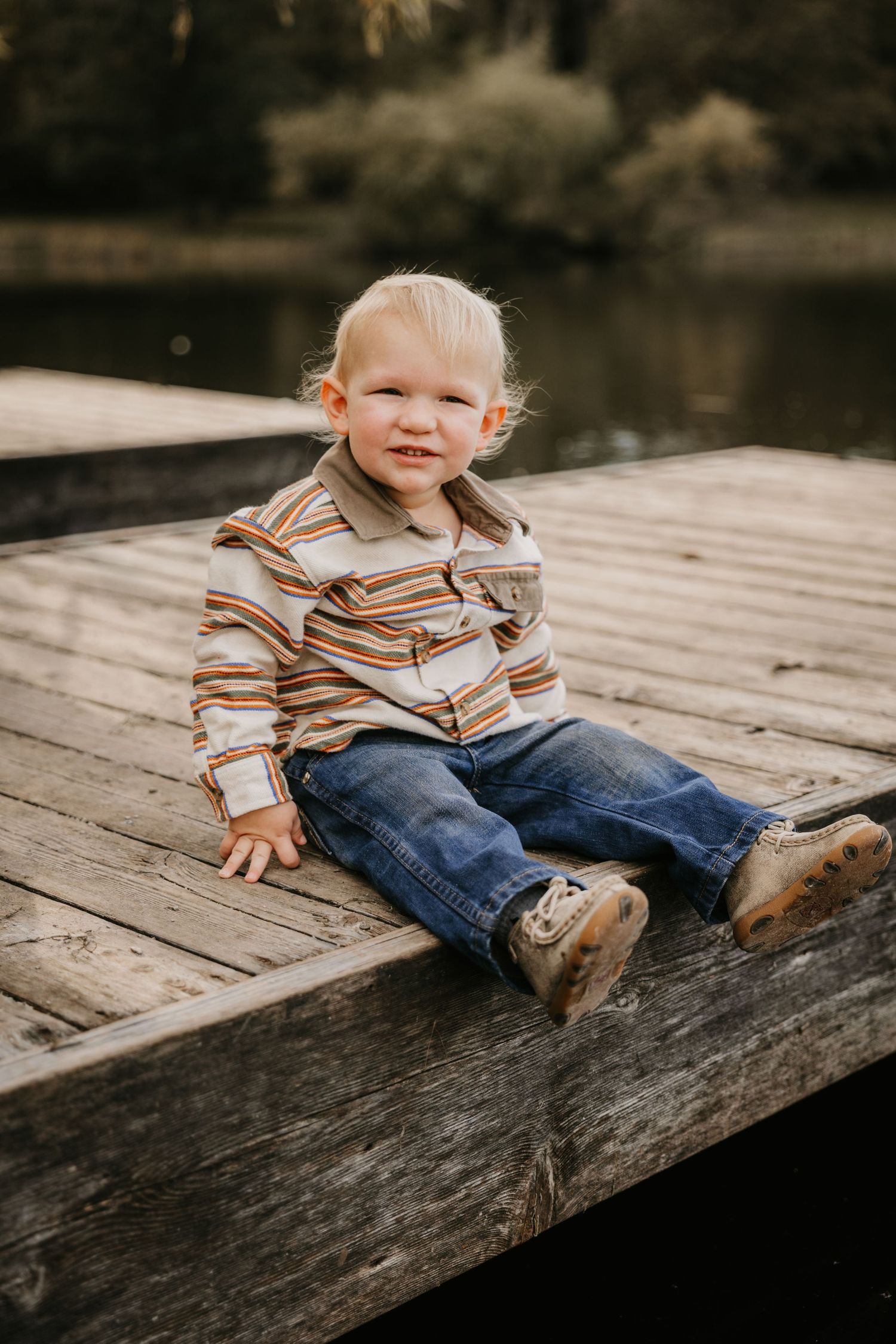 Smiling blond toddler in a striped western shirt and boots sits on a weathered wooden dock beside a calm lake.