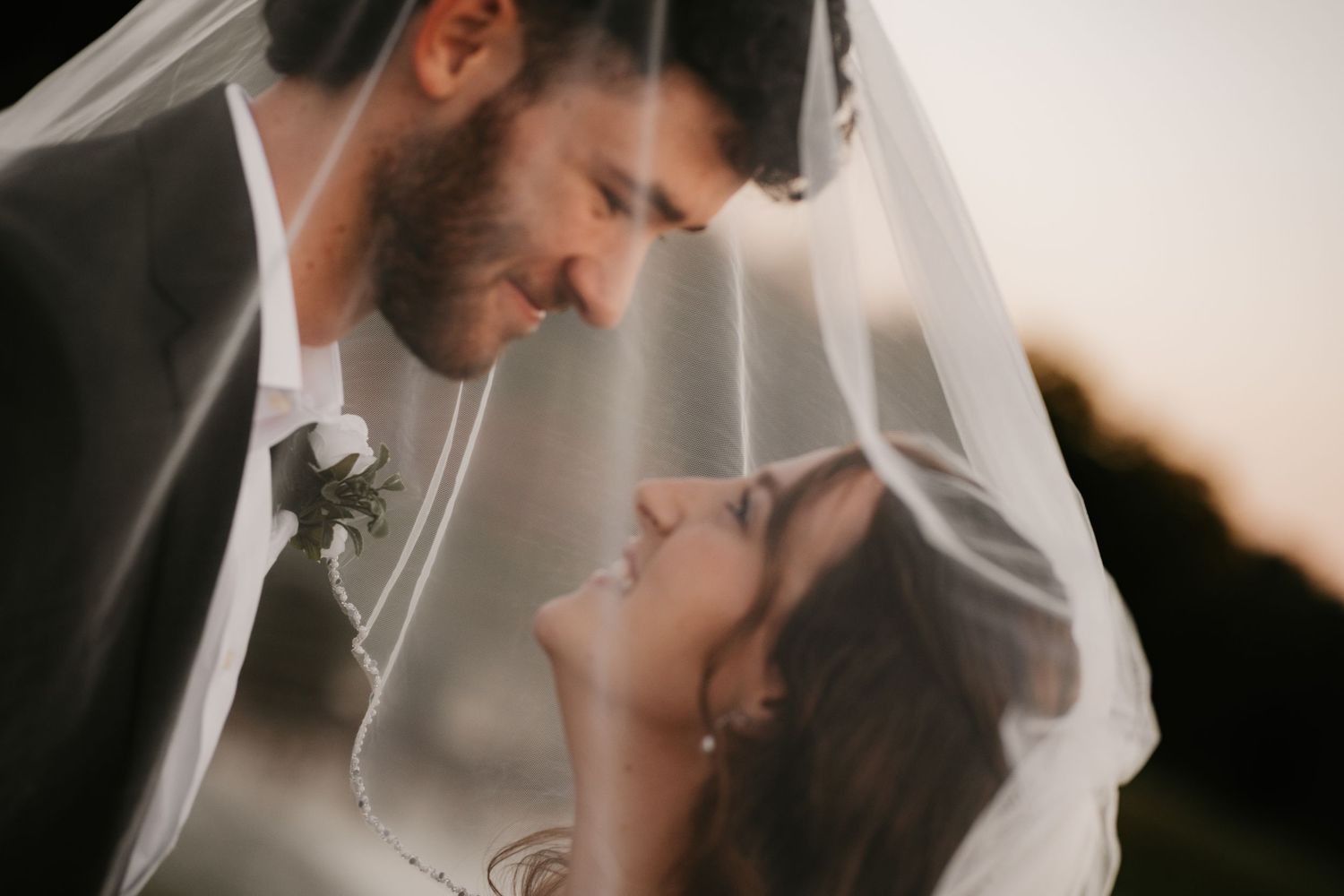 Groom with a boutonniere leans in to kiss his bride beneath her sheer wedding veil in warm sunset light.