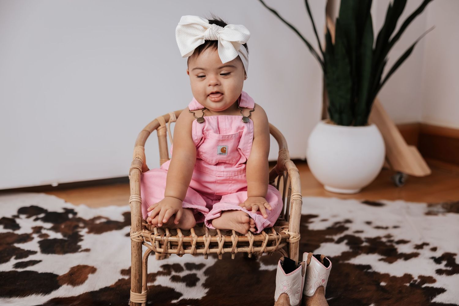 Baby girl in pink overalls and a white bow headband sits in a rattan chair on a cowhide rug beside a potted plant.