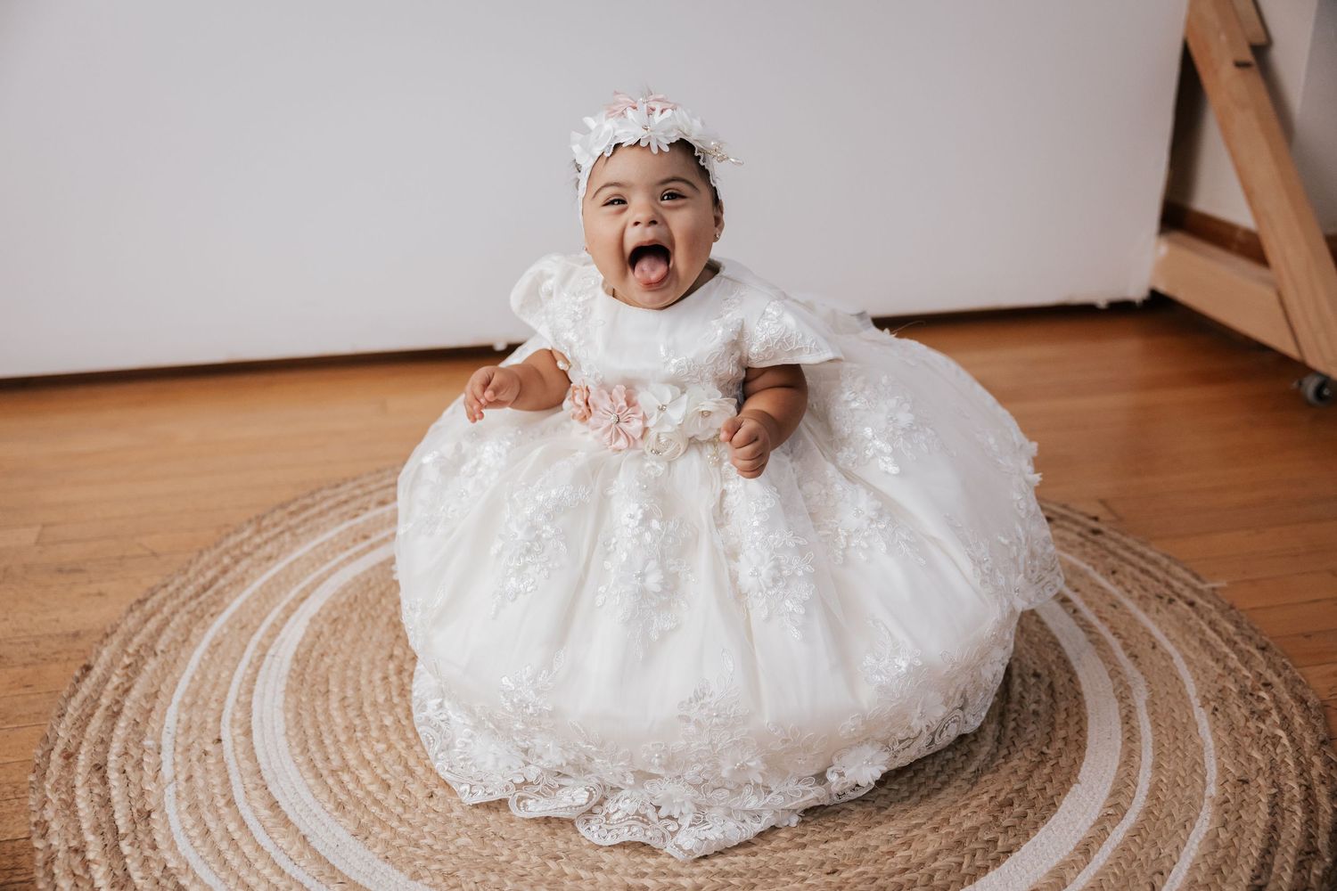 Laughing baby in a white lace christening gown and floral headband sits on a round jute rug in a bright studio.