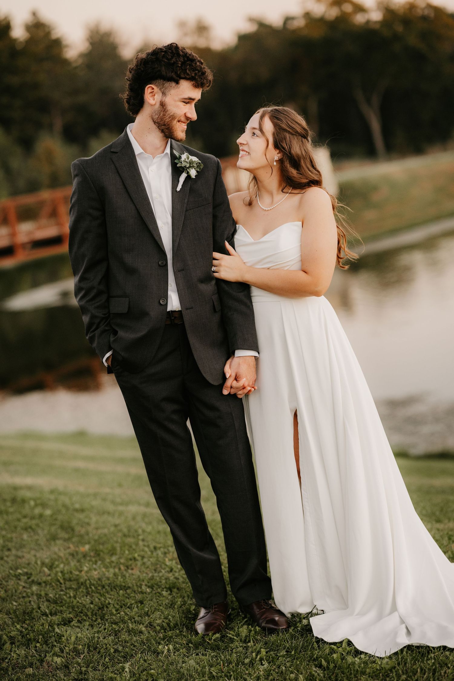 Smiling groom in a dark suit holds hands with his bride in a white gown with a leg slit while standing on a grassy lawn by a pond.