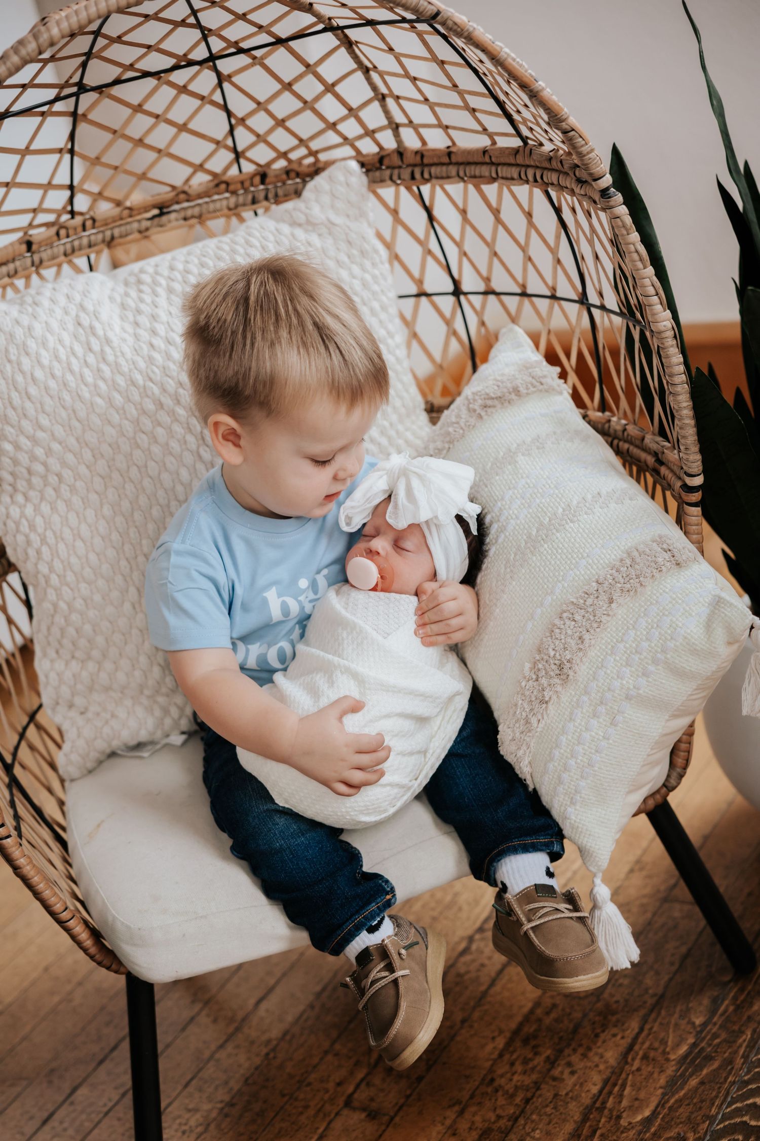 Big brother in a light blue shirt and jeans cradles his sleeping newborn sister with a pacifier in a rattan chair with pillows.