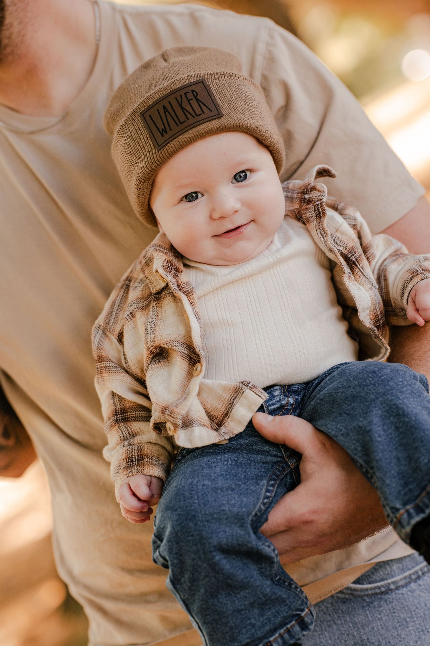 Smiling baby with blue eyes wears a tan Walker beanie and plaid flannel shirt while held outdoors by a parent.