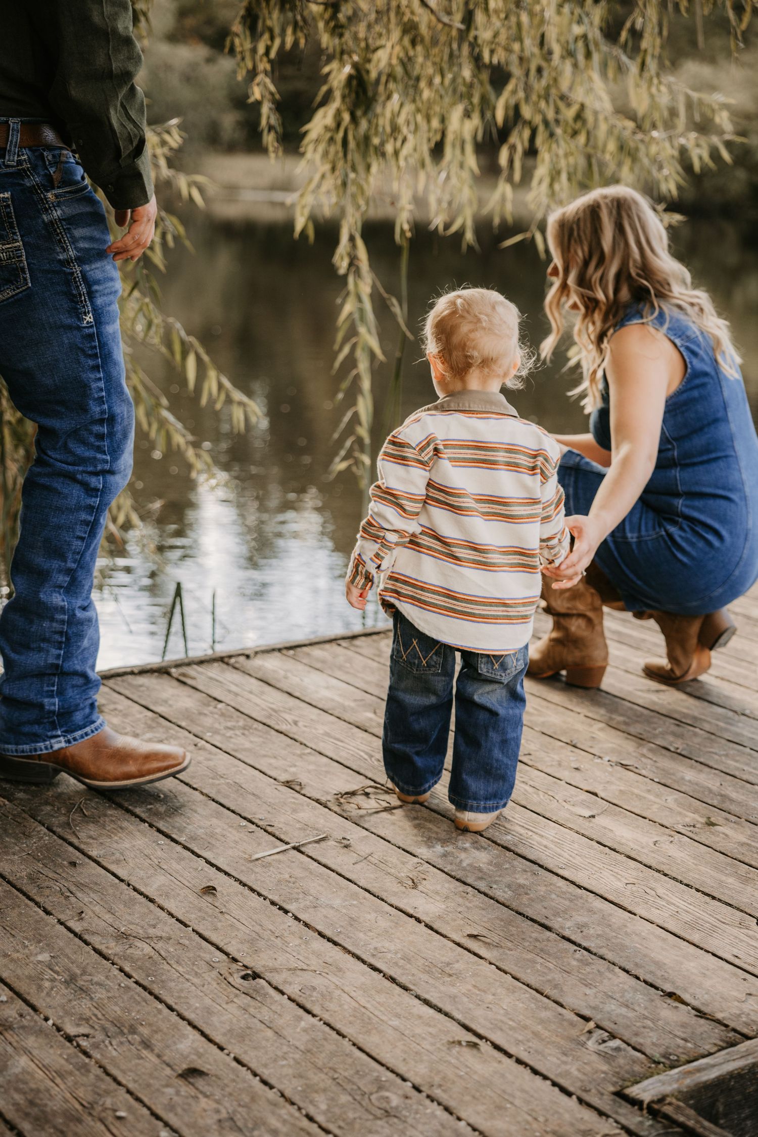 Toddler in a striped shirt stands on a wooden dock with his parents, looking out over a peaceful lake framed by willow branches.