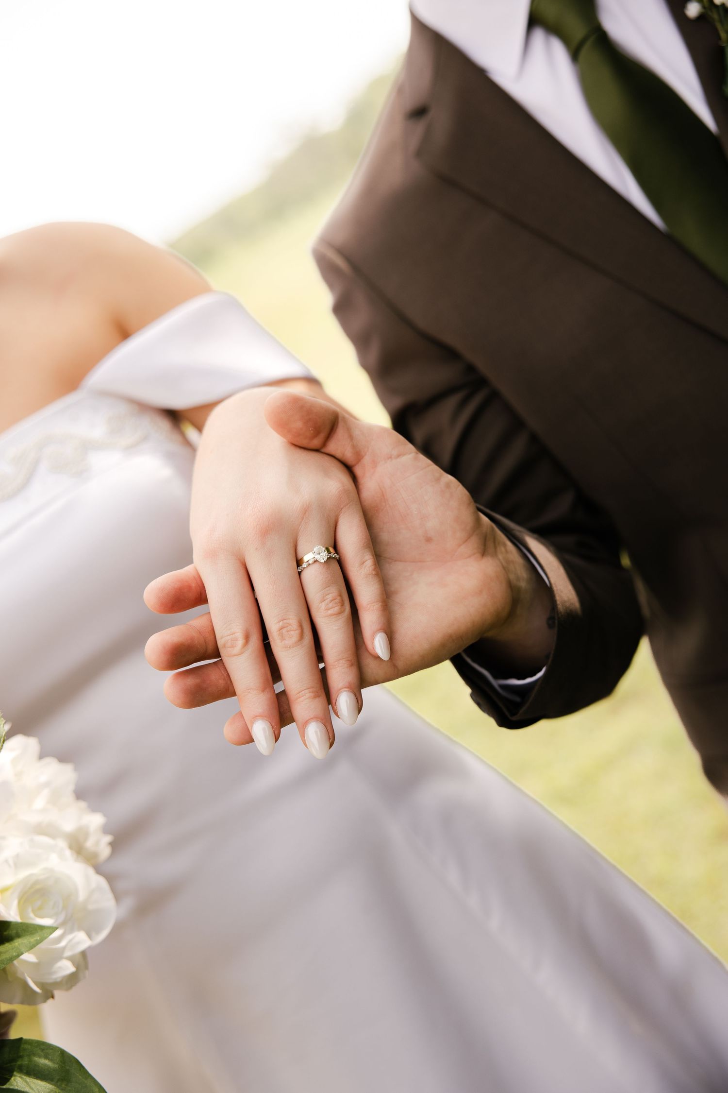 Close-up of a bride and groom holding hands, showing a diamond engagement ring and wedding band against a white dress and brown suit.