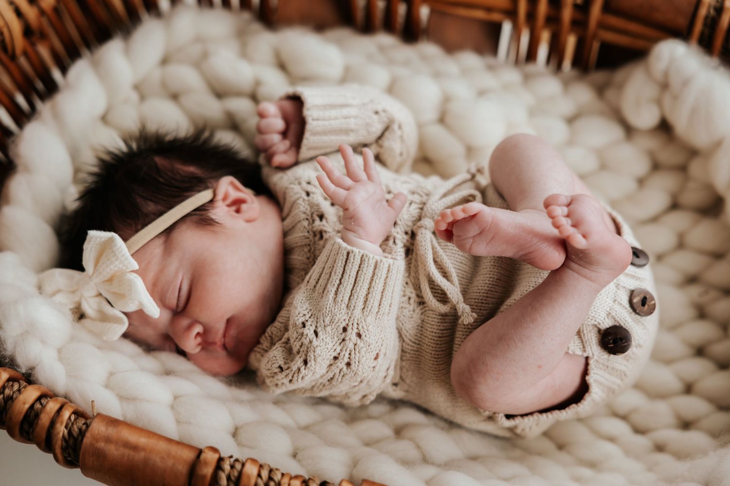 Peaceful newborn in a knit cream romper and bow headband sleeps in a cozy basket on a soft chunky knit blanket.