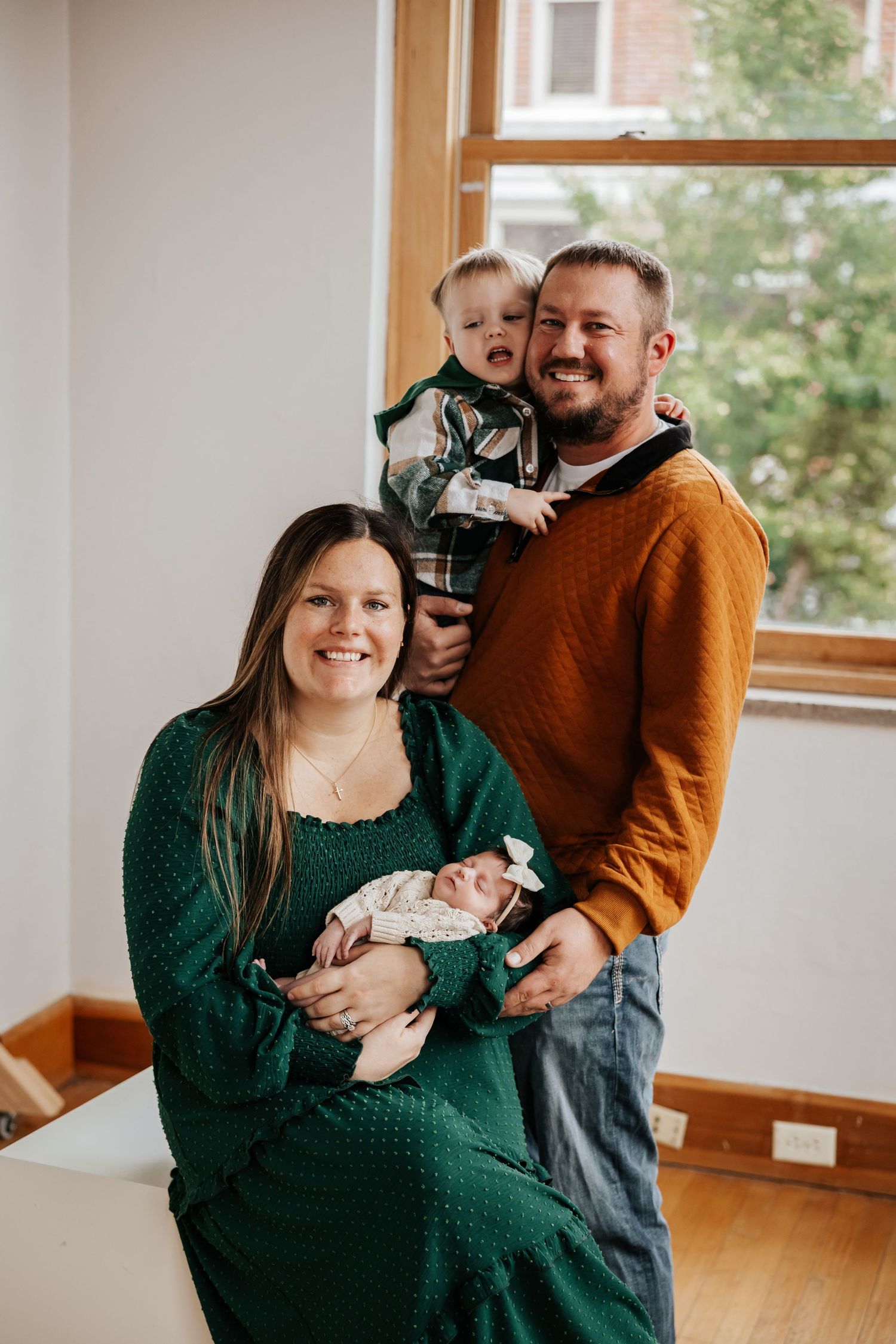 A happy family portrait shows a smiling mother cradling her newborn while a father holds a cheerful toddler by the window.
