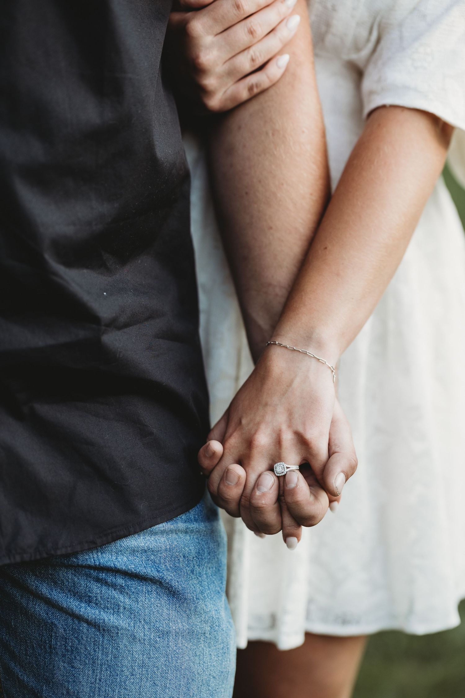 Couple holding hands in close-up, showing a sparkling engagement ring, with a white lace dress and denim jeans.