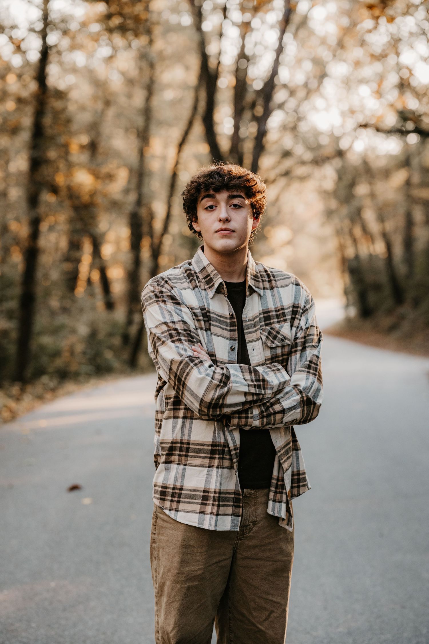 Teenage boy with curly hair and arms crossed stands in a plaid flannel shirt on a tree-lined country road in autumn.