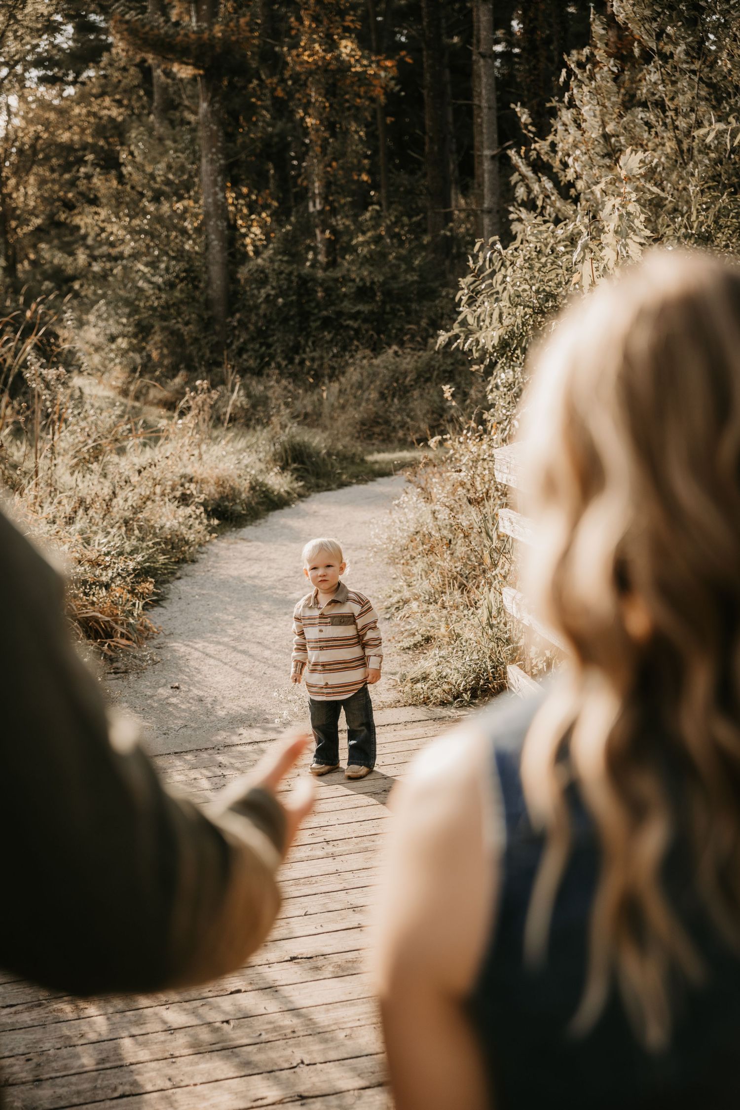 A toddler in a striped sweater stands on a wooden boardwalk path through the woods as two adults reach toward him.