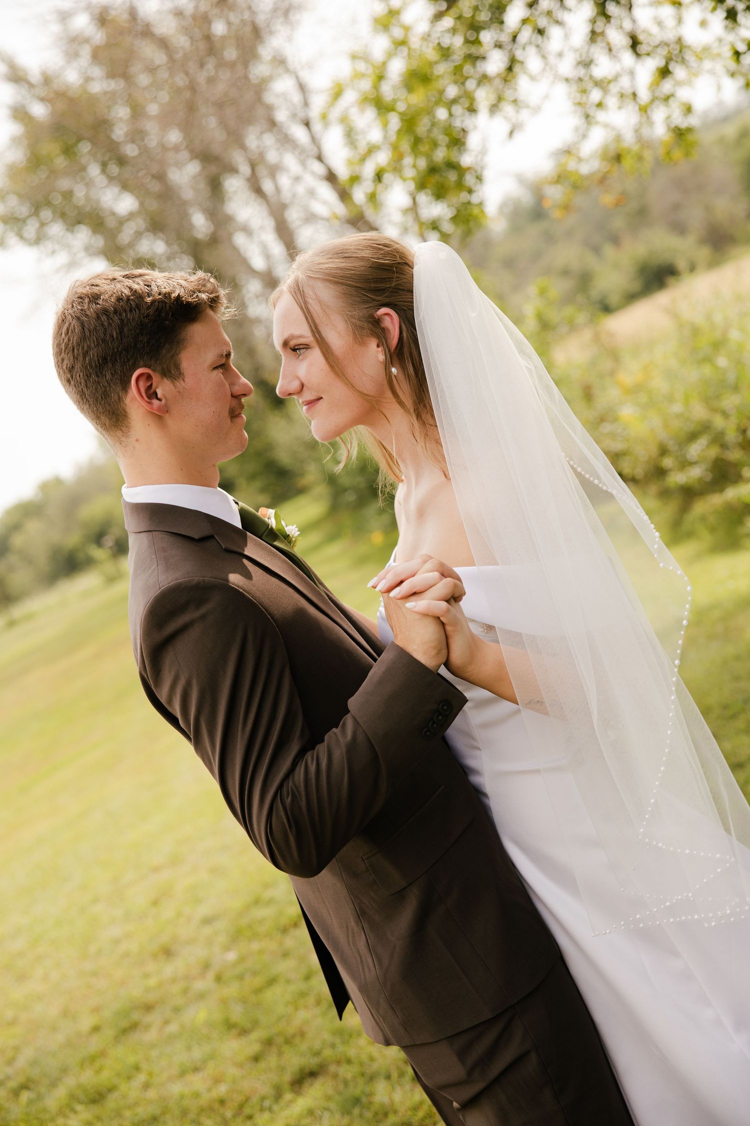 A bride in a long beaded veil and a groom in a brown suit gaze at each other while holding hands in a green, sunlit outdoor field.