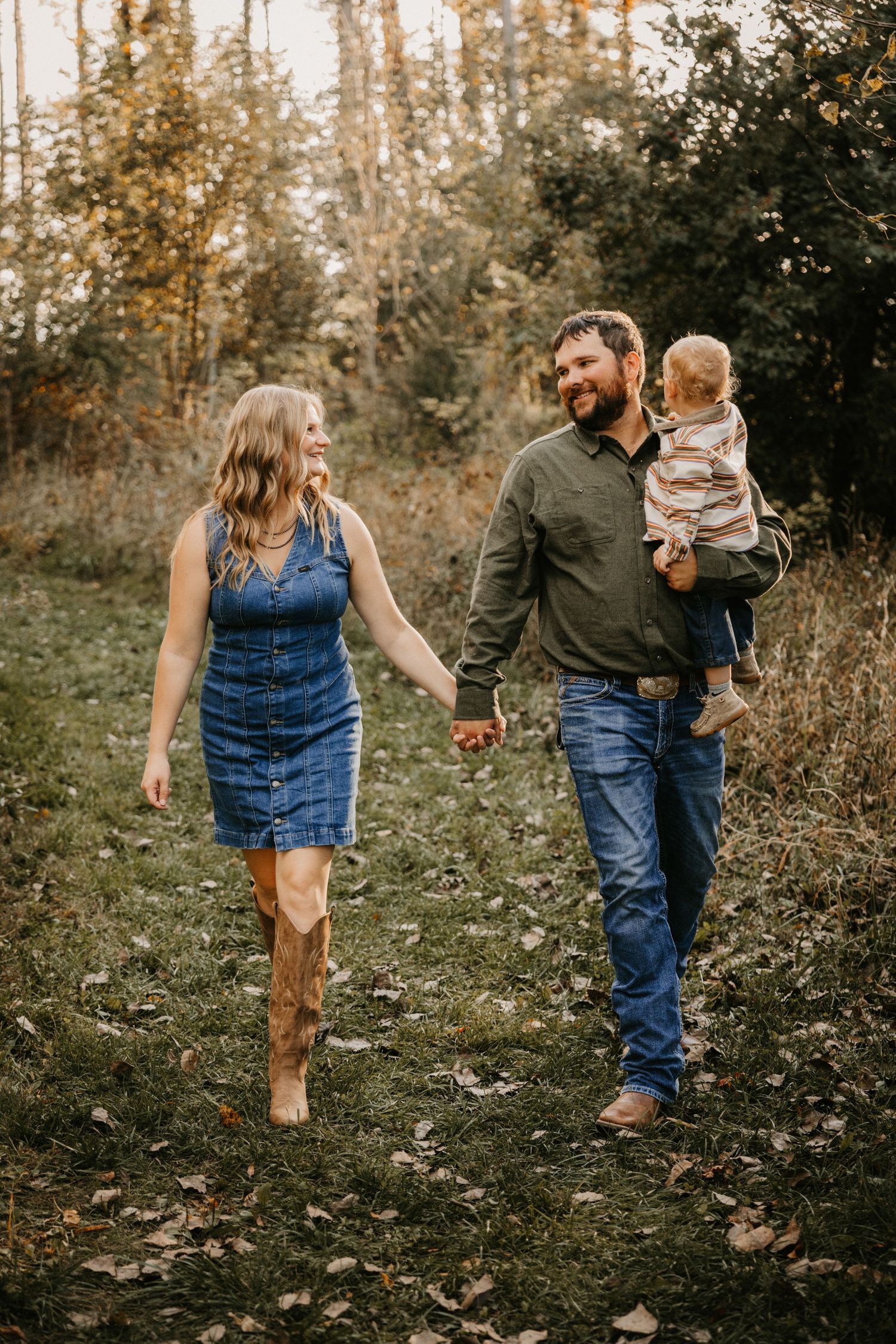 A woman in a denim dress and cowboy boots smiles at her husband as he carries their toddler on a leaf-covered trail.