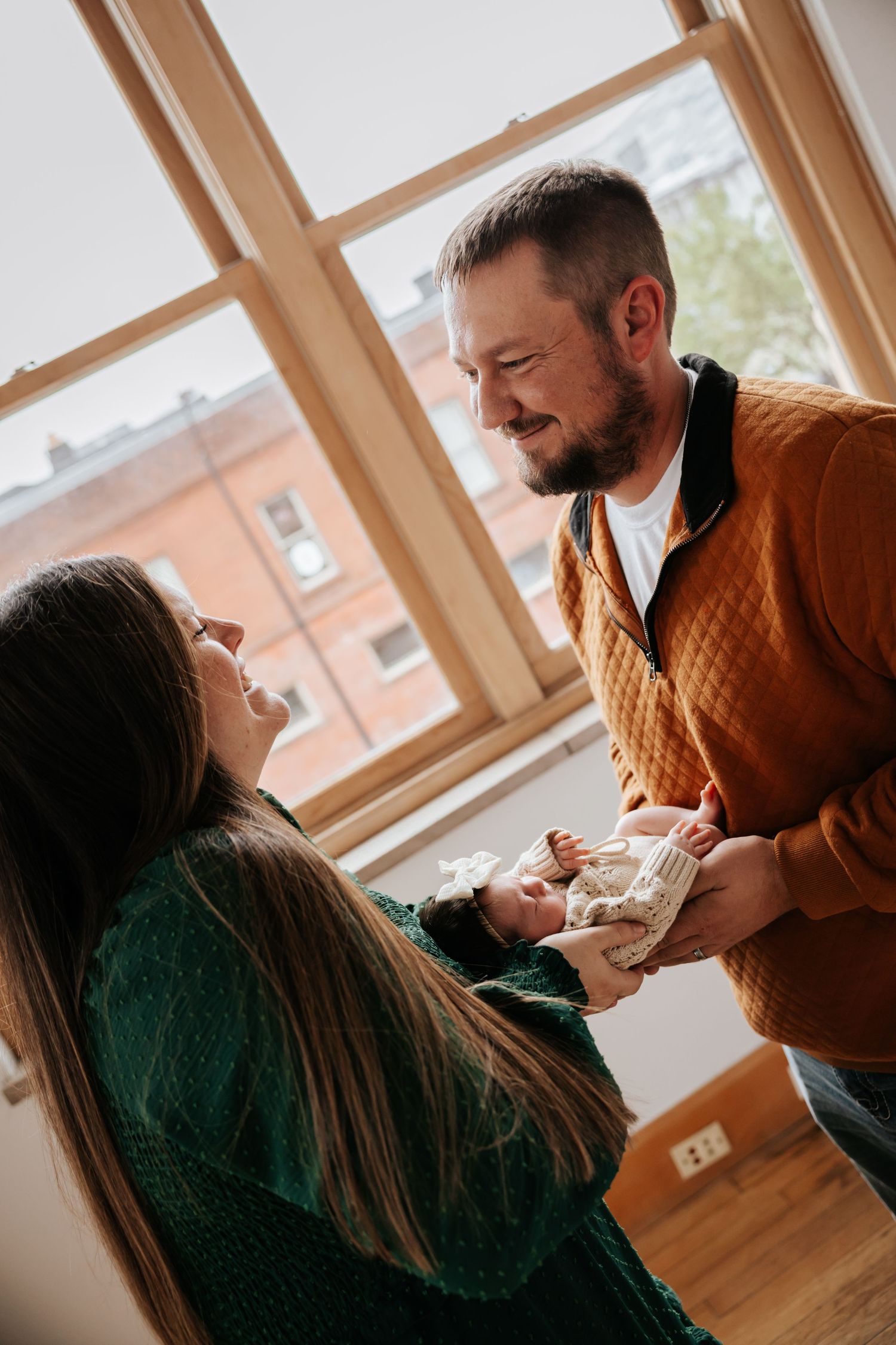 Smiling parents hold their newborn baby between them in a bright room with large windows and warm wood floors.