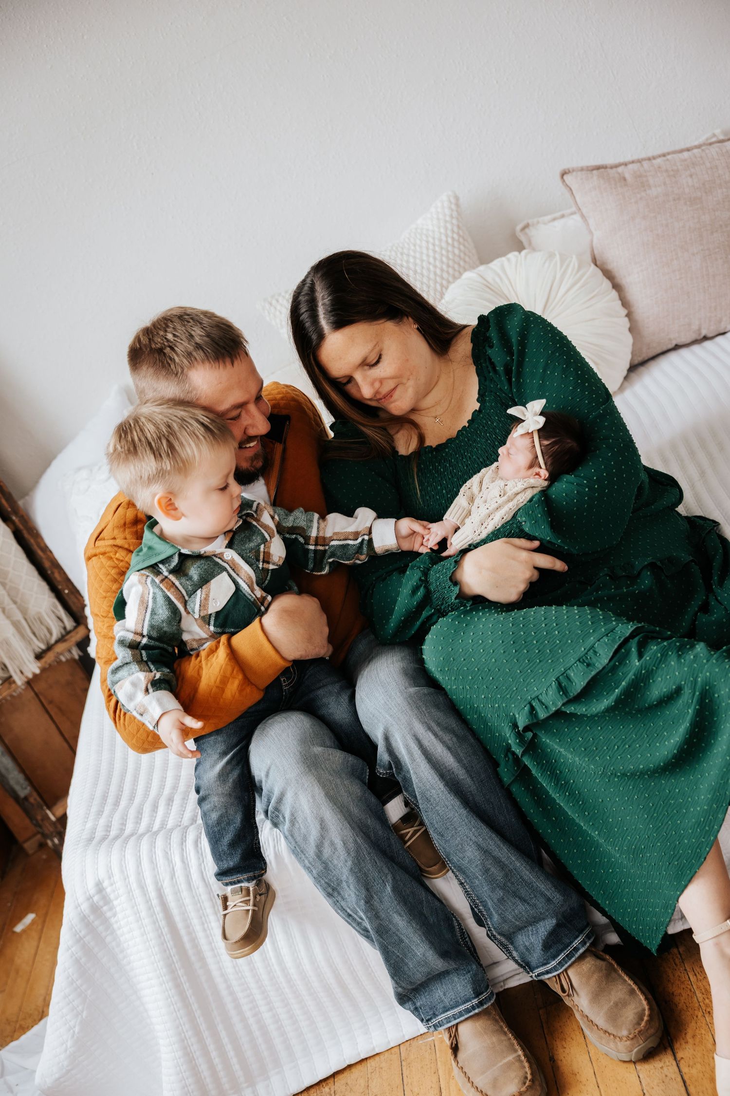 An overhead view of a family on a daybed as a toddler reaches toward a newborn baby cradled in the mother's arms.
