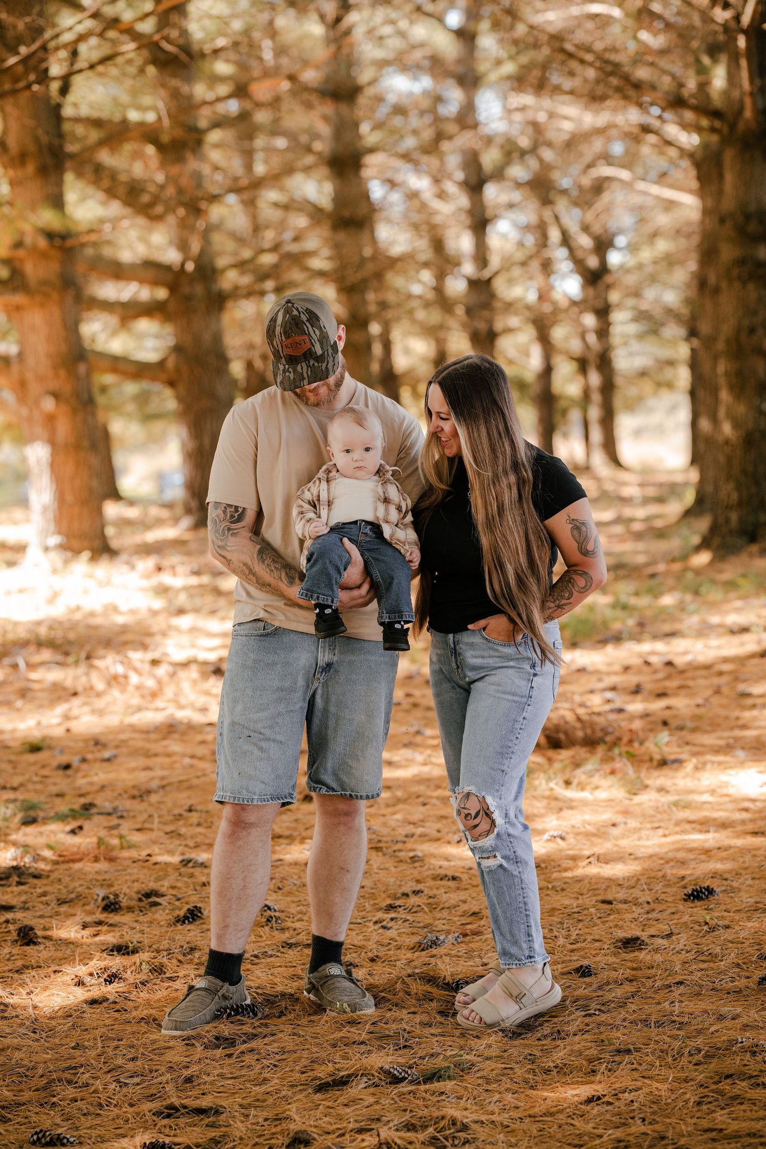 Tattooed couple stands on a pine needle forest floor and smiles down at their baby in a plaid shirt during a family photo session.
