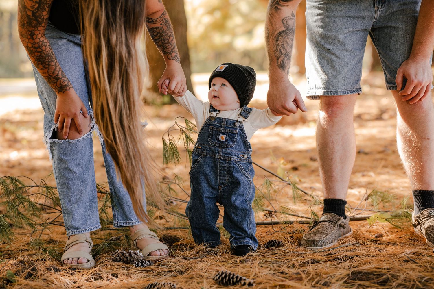 Tattooed parents hold the hands of a toddler in overalls and a beanie as the child takes steps on pine needles and pinecones.