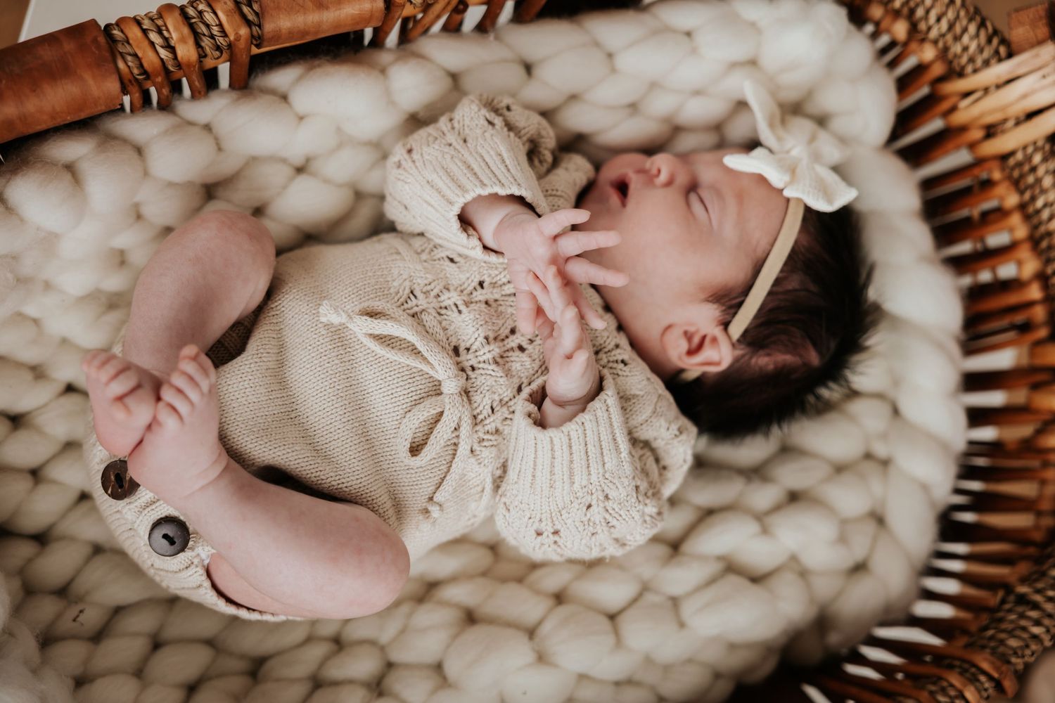 Sleeping newborn girl in a knit cream outfit and bow headband rests in a woven basket lined with chunky wool.