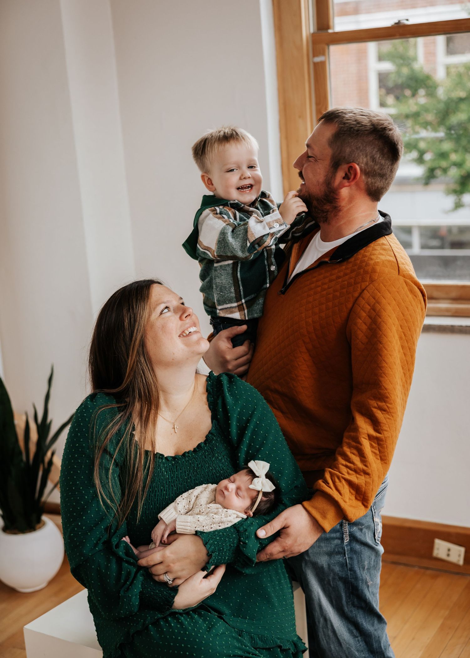 A mother smiles lovingly at her newborn as a father holds a laughing toddler high in a cozy indoor family photo session.