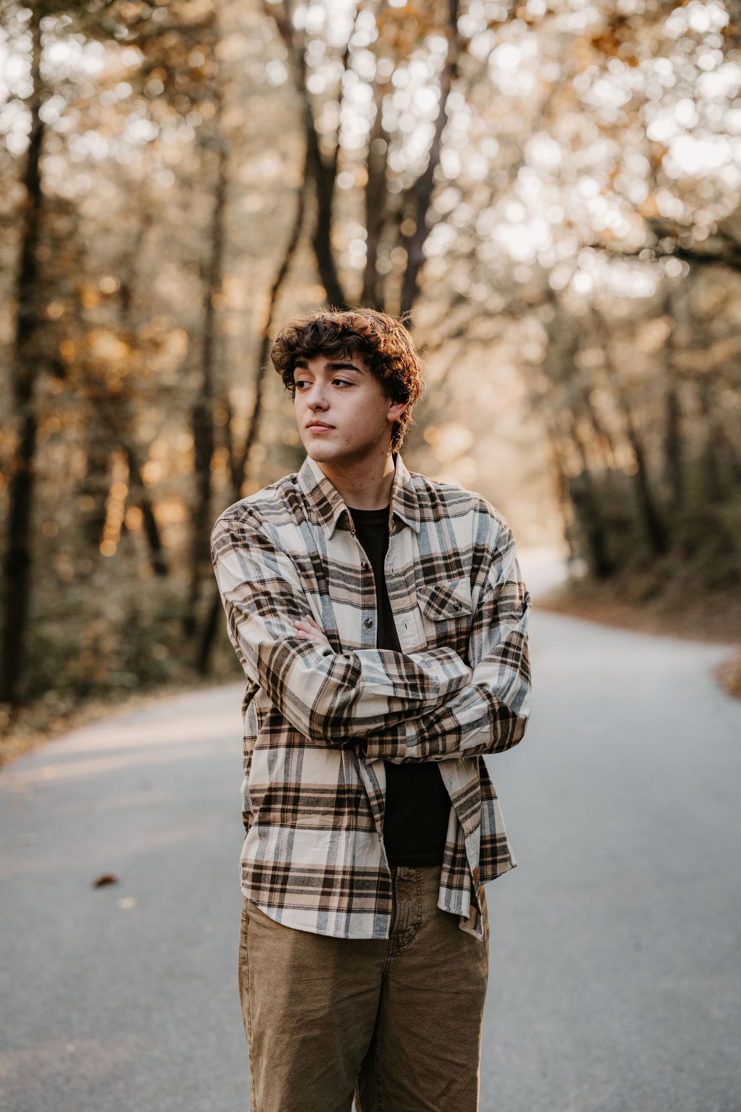 Young man in a plaid flannel shirt and khaki pants stands with arms crossed on a country road lined with autumn trees.