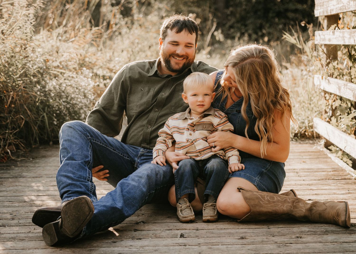 A happy family sits on a rustic wooden bridge surrounded by tall grass, with the mom in cowboy boots hugging her toddler son and husband.