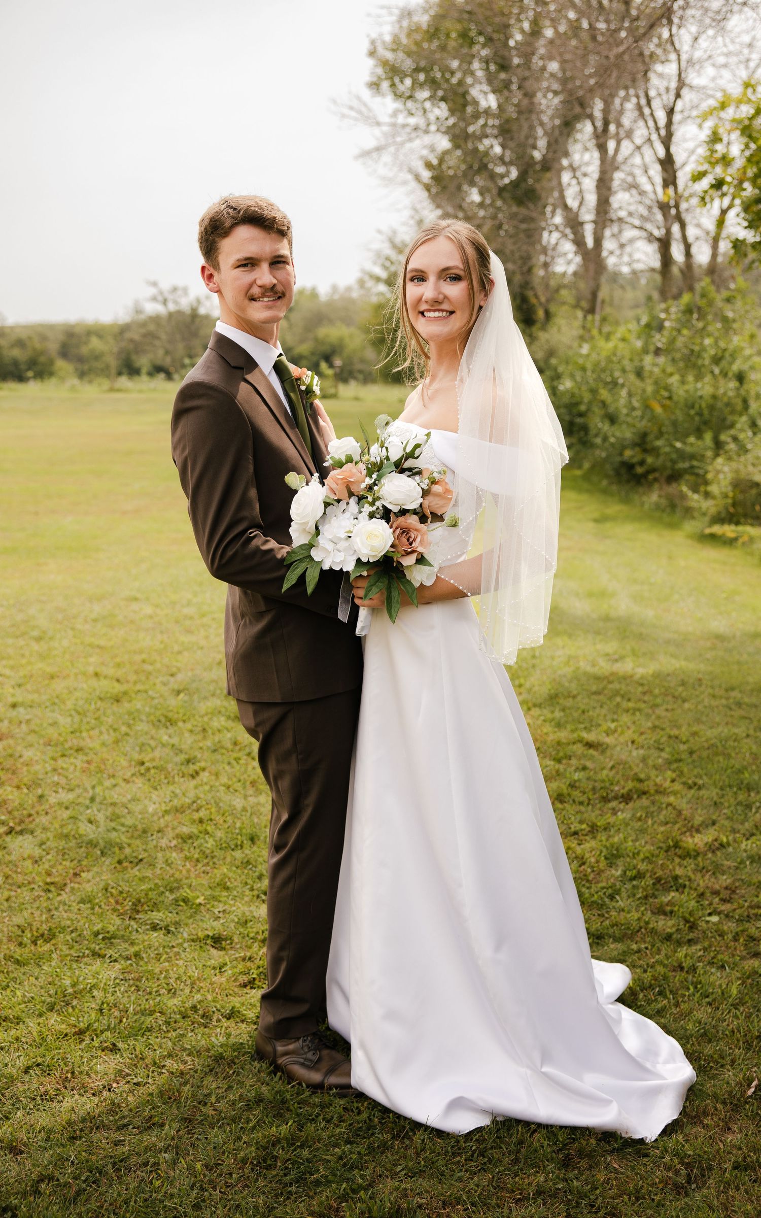 A smiling bride in a white satin gown and veil poses with a groom in a brown suit, holding a white and peach bouquet on a grassy lawn.