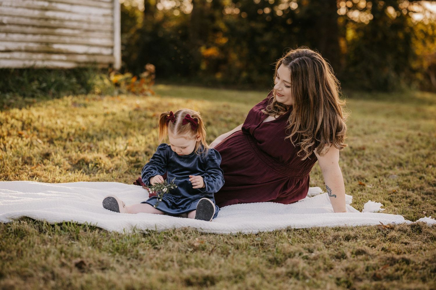 Pregnant mother in a burgundy dress sits on a white blanket in the grass beside her toddler daughter during a family portrait session.