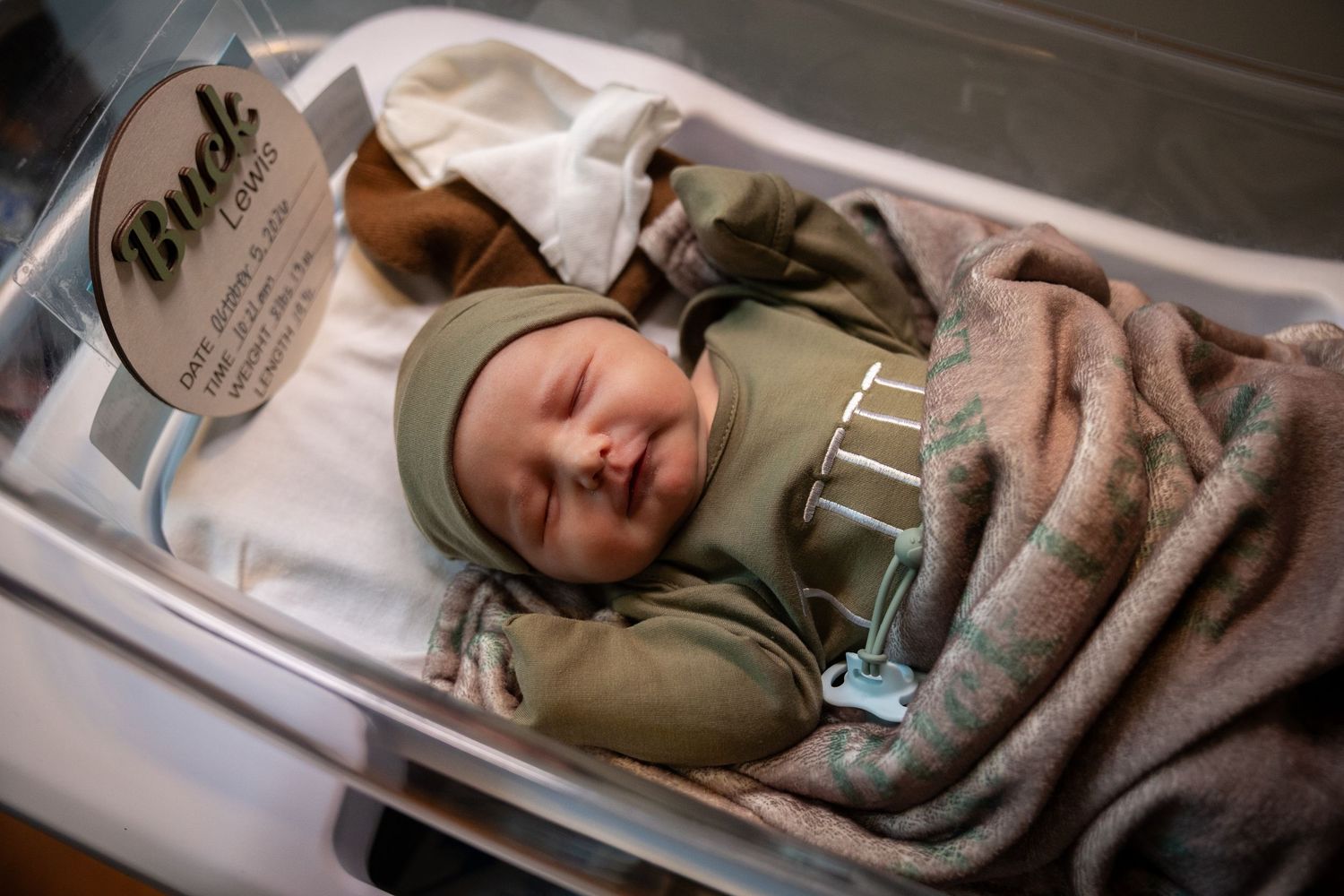A newborn baby in an olive green outfit and hat sleeps peacefully in a clear hospital bassinet with a name card attached.