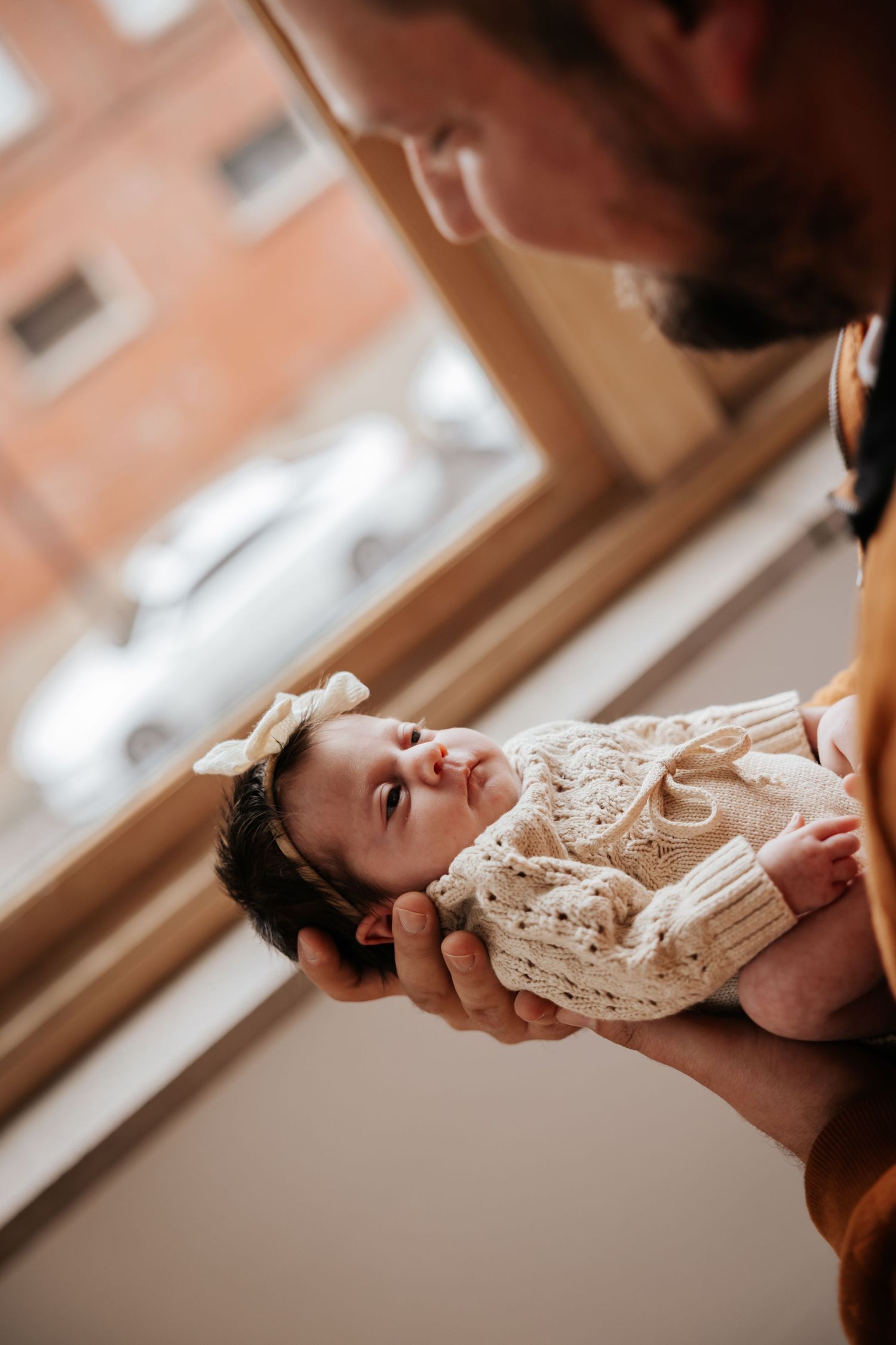 Newborn baby in a cream knit romper and bow headband looks up while resting in a father's hands near a window.
