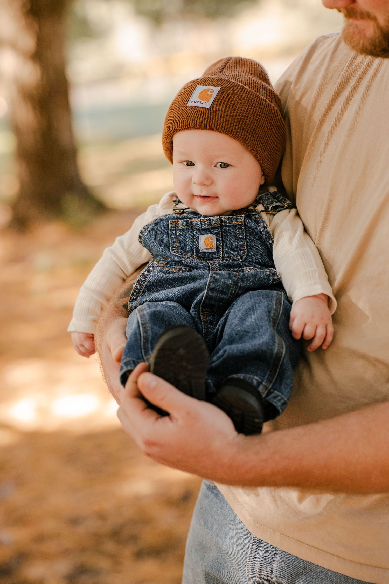 Smiling baby in a brown Carhartt beanie and denim overalls is held by a bearded parent in a sunlit autumn forest.