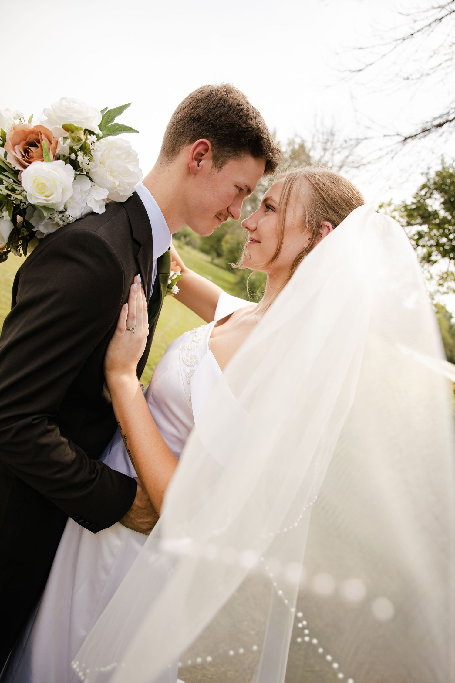 Groom leans close to his bride as she wears a beaded veil and holds a white and peach rose bouquet.