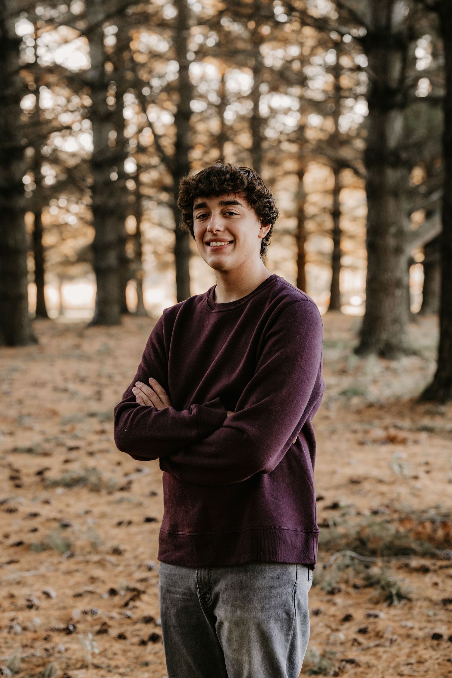 Smiling young man with curly hair stands with arms crossed in a maroon sweatshirt among tall pine trees in warm golden light.