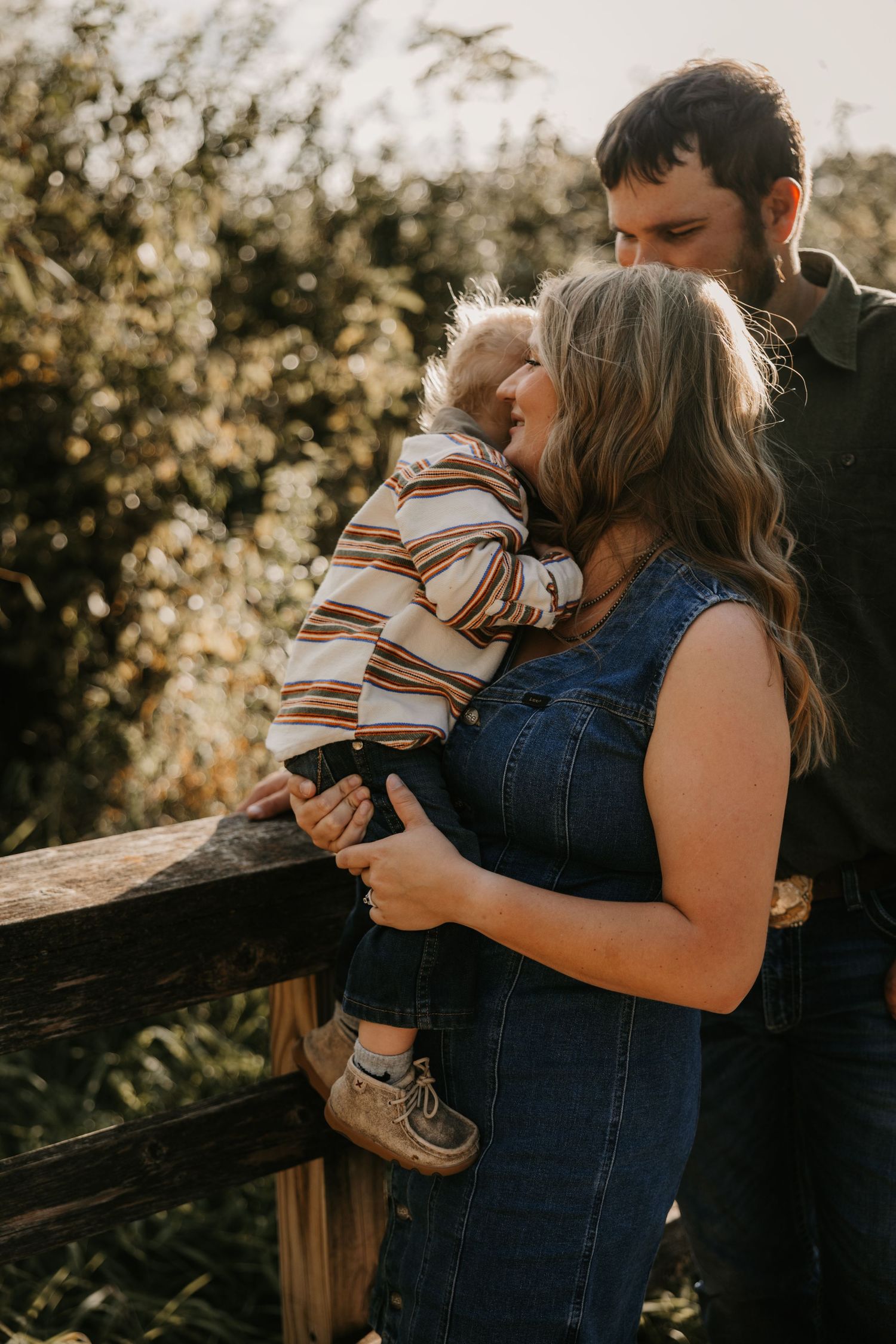 A mother in a denim dress cuddles her toddler against a rustic wooden fence while her husband stands close by in warm evening light.