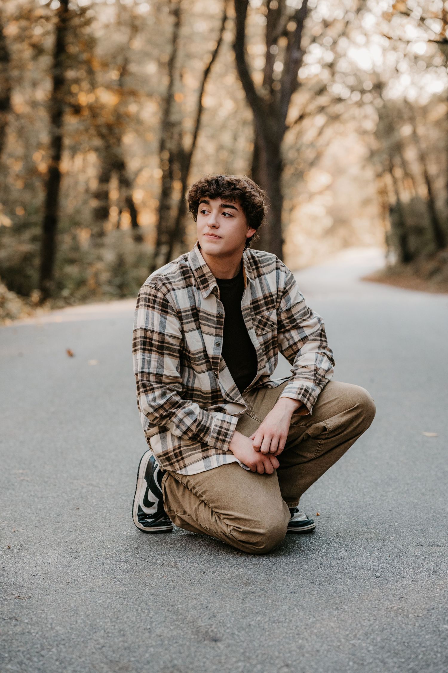 Young man in a plaid flannel shirt and khaki pants crouches on a paved forest road, looking off to the side in autumn light.