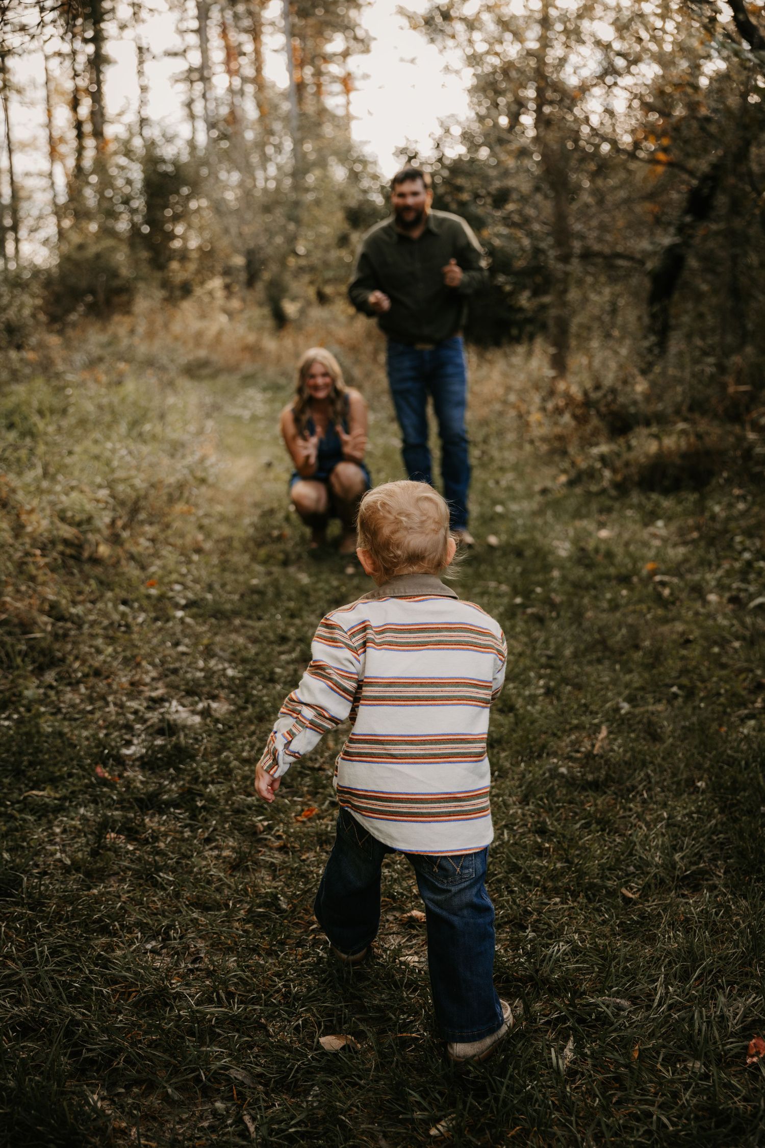A toddler in a striped shirt walks down a wooded path toward his smiling parents as they crouch and reach out to greet him.