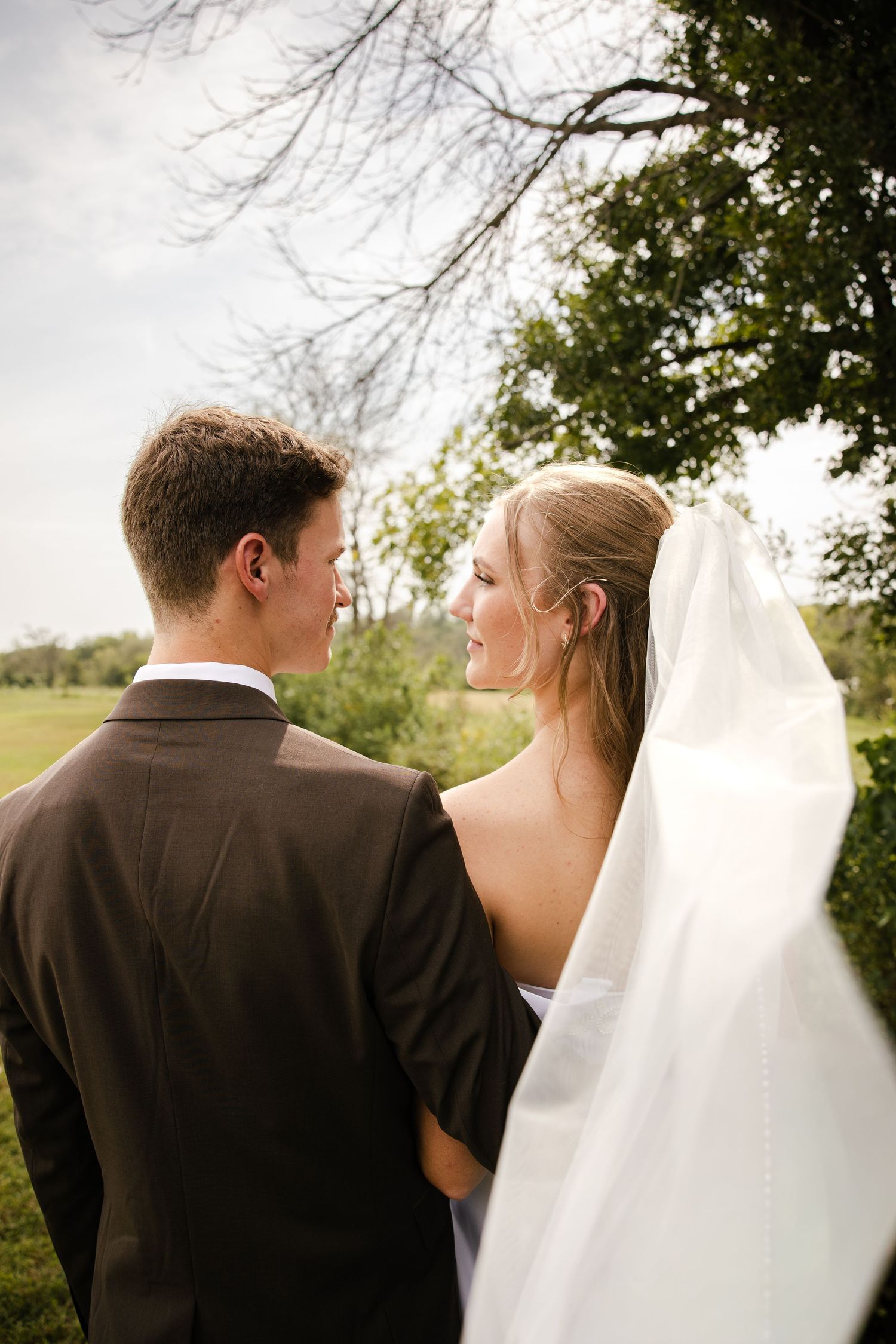 Groom in a brown suit gazes at his bride in a long white veil beneath green trees on their wedding day.