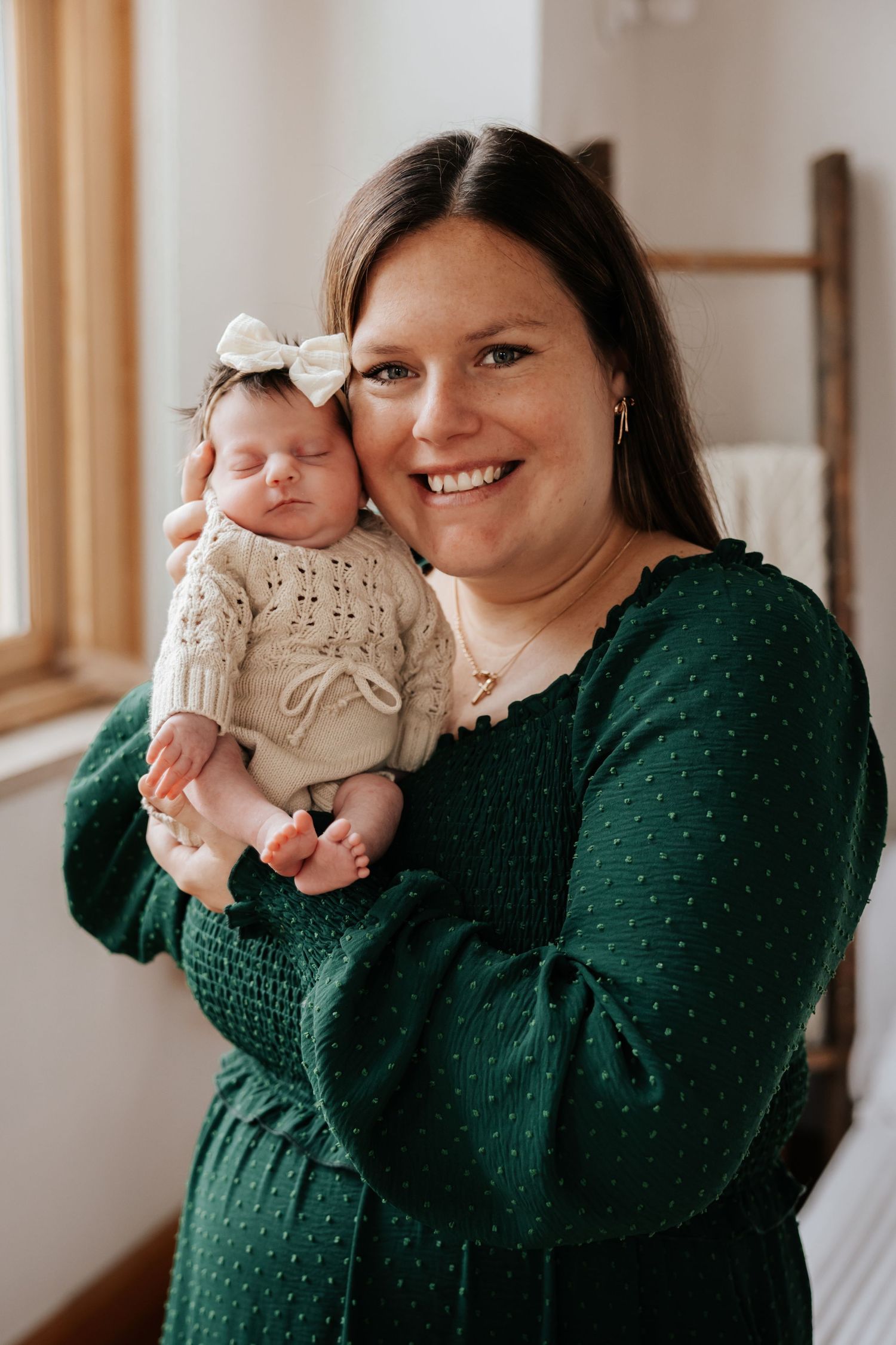 Happy mom in an emerald green dress holds her sleeping newborn baby girl in a cream knit outfit near a window.