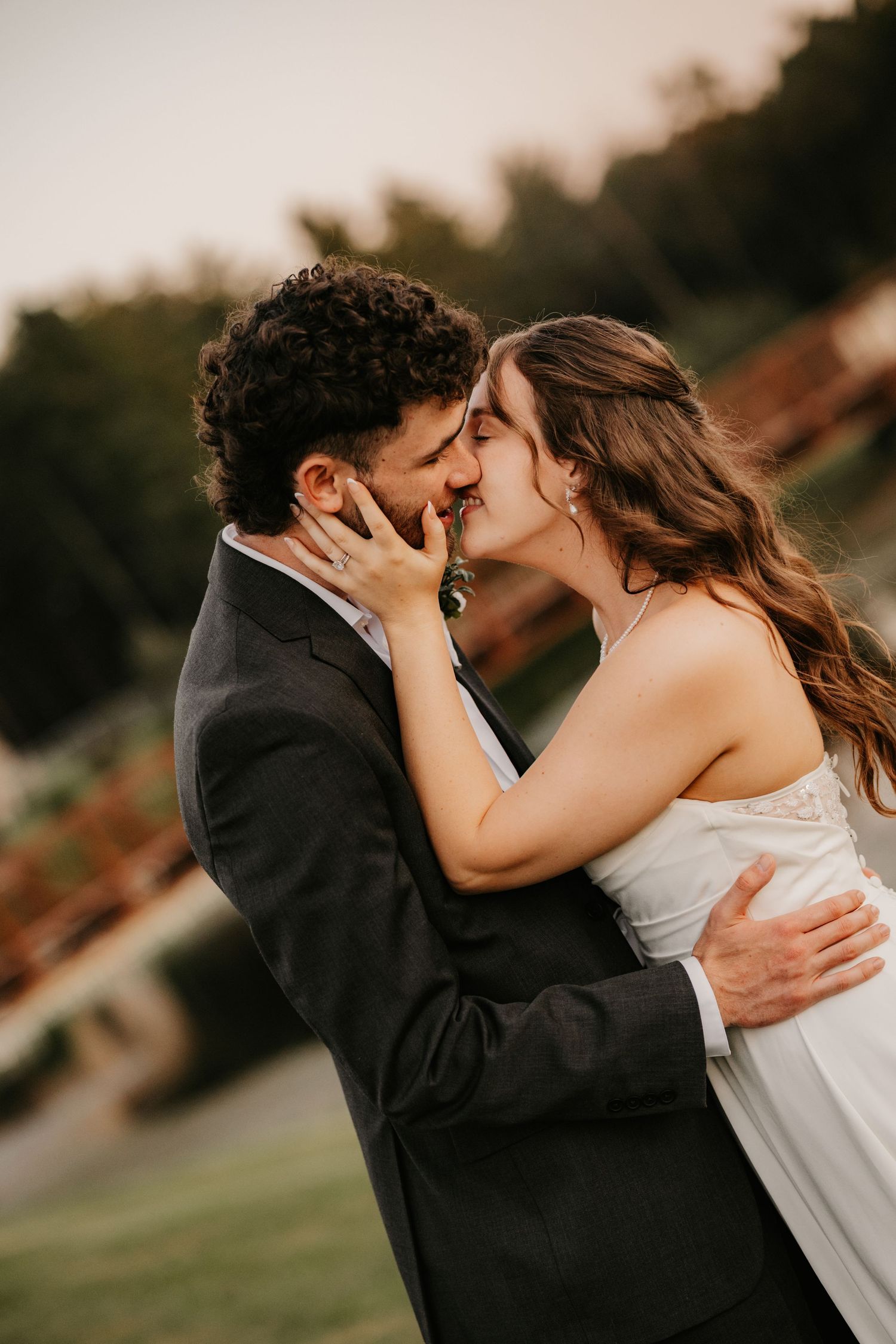 Bride and groom share a romantic kiss outdoors at golden hour, her hand on his cheek and his arm around her waist.