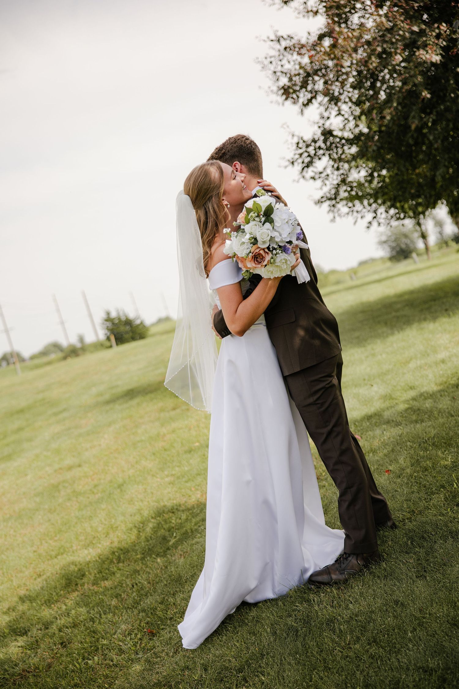 Bride and groom embrace in a tender hug on a grassy lawn, the bride holding a white and blue bouquet with her veil flowing behind.