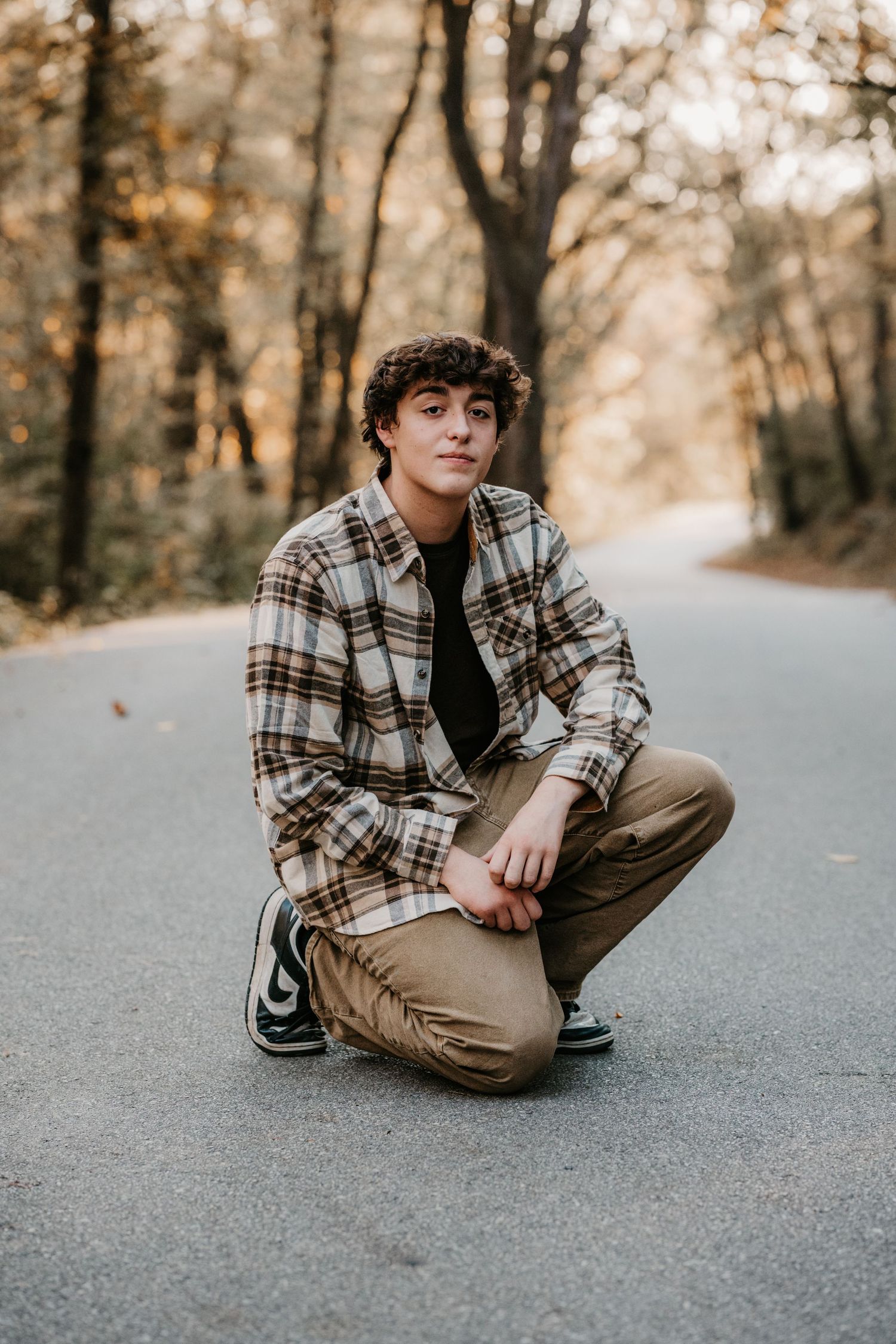 Young man with curly hair crouching on a tree-lined road in a plaid flannel shirt, tan pants and sneakers during golden hour.