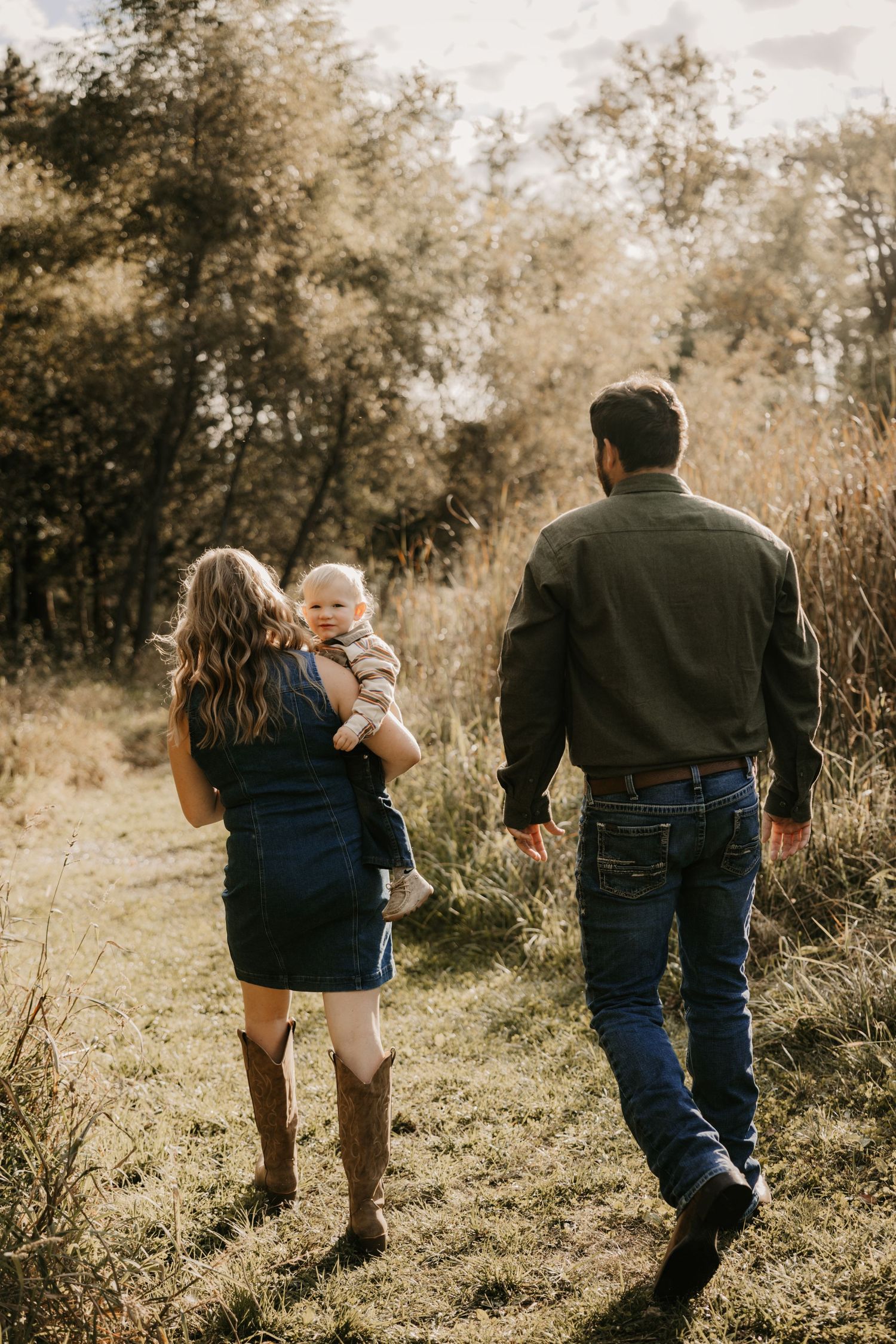 Parents in western clothing walk away through a grassy field, mother carrying the smiling toddler over her shoulder in warm sunlight.